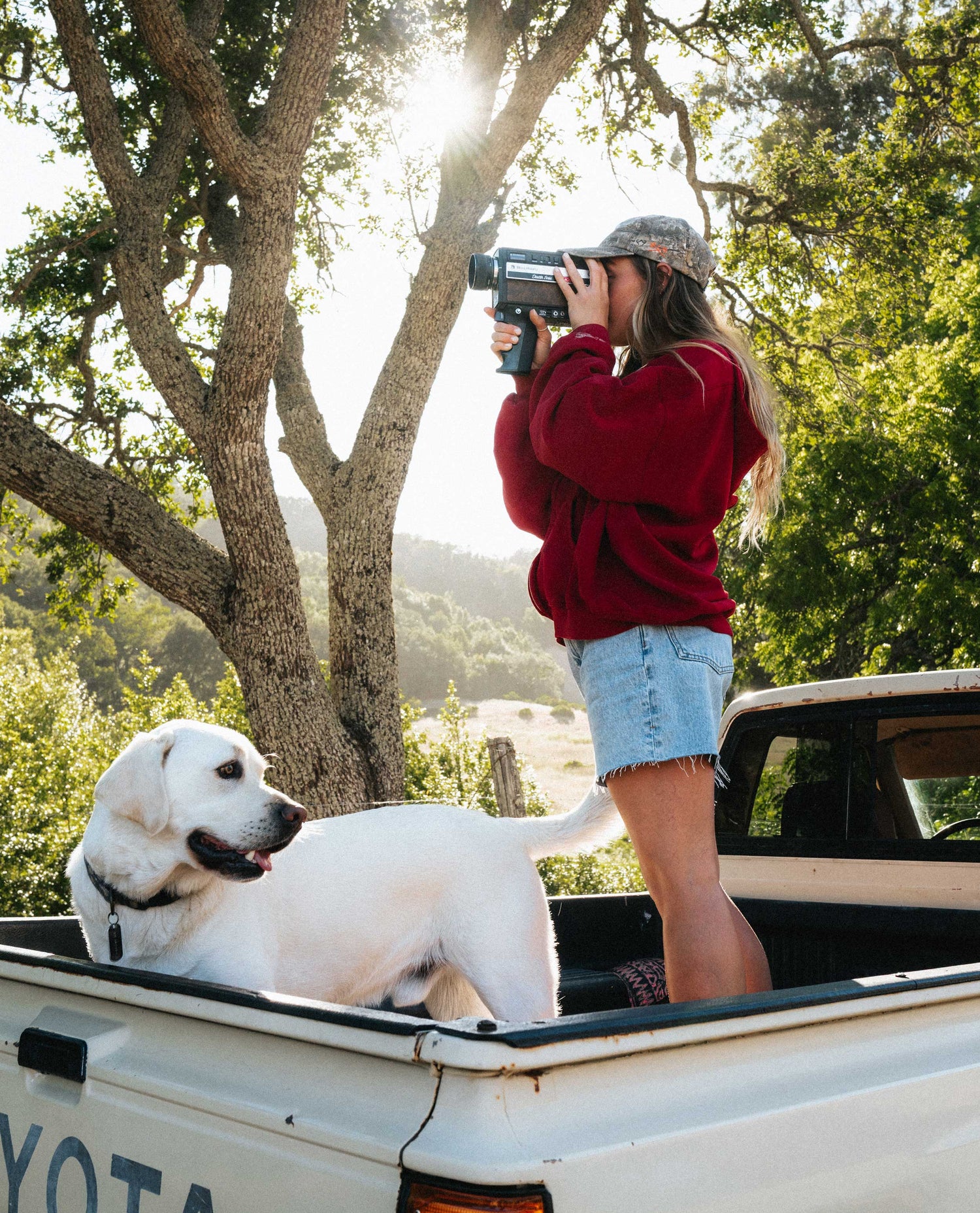 Girl wearing ight blue denim shorts and red top, holding an old camera and standing with a dog in the bed of a truck.