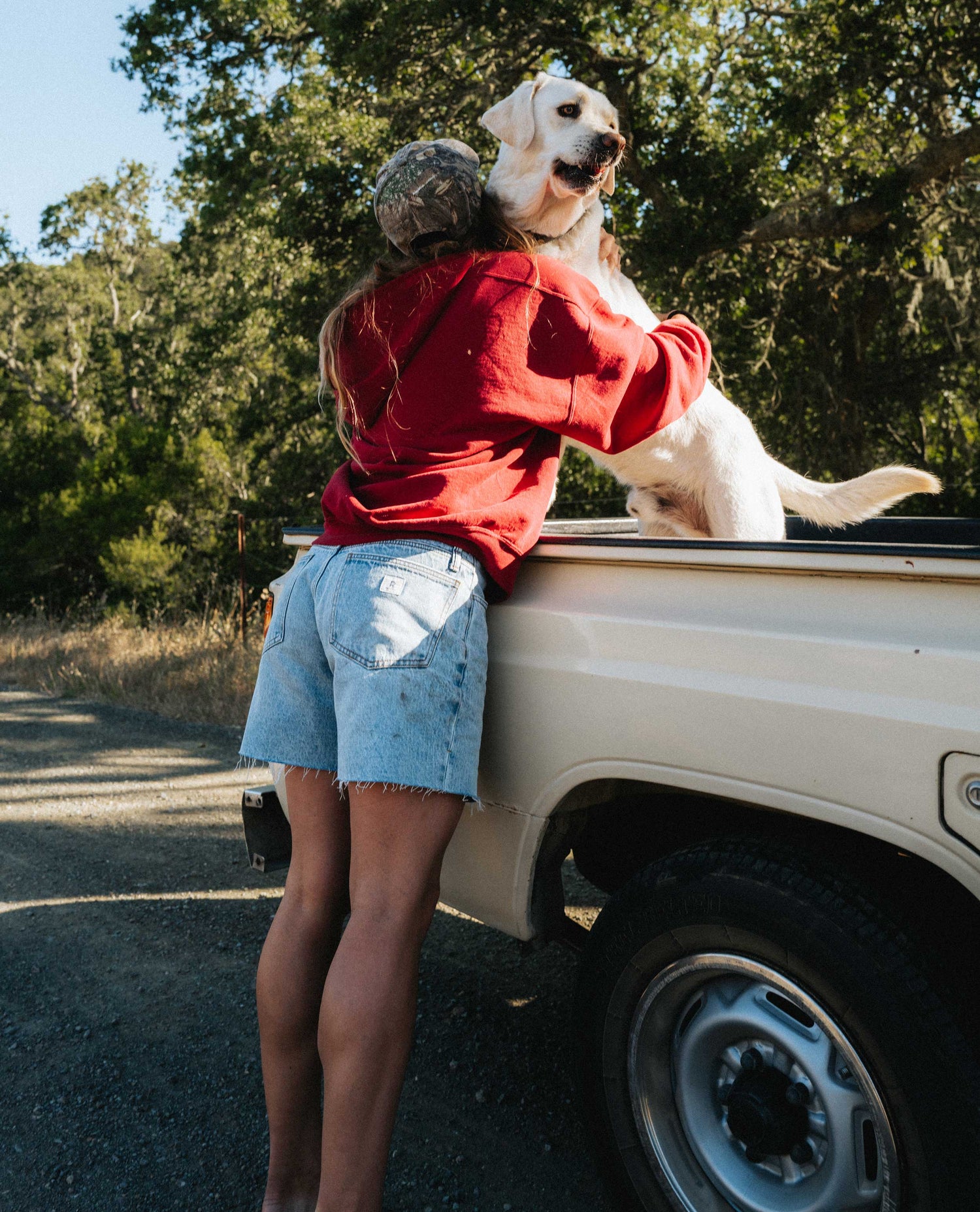 Girl wearing light blue denim shorts and red shirt, hugging a dog in a truck. 