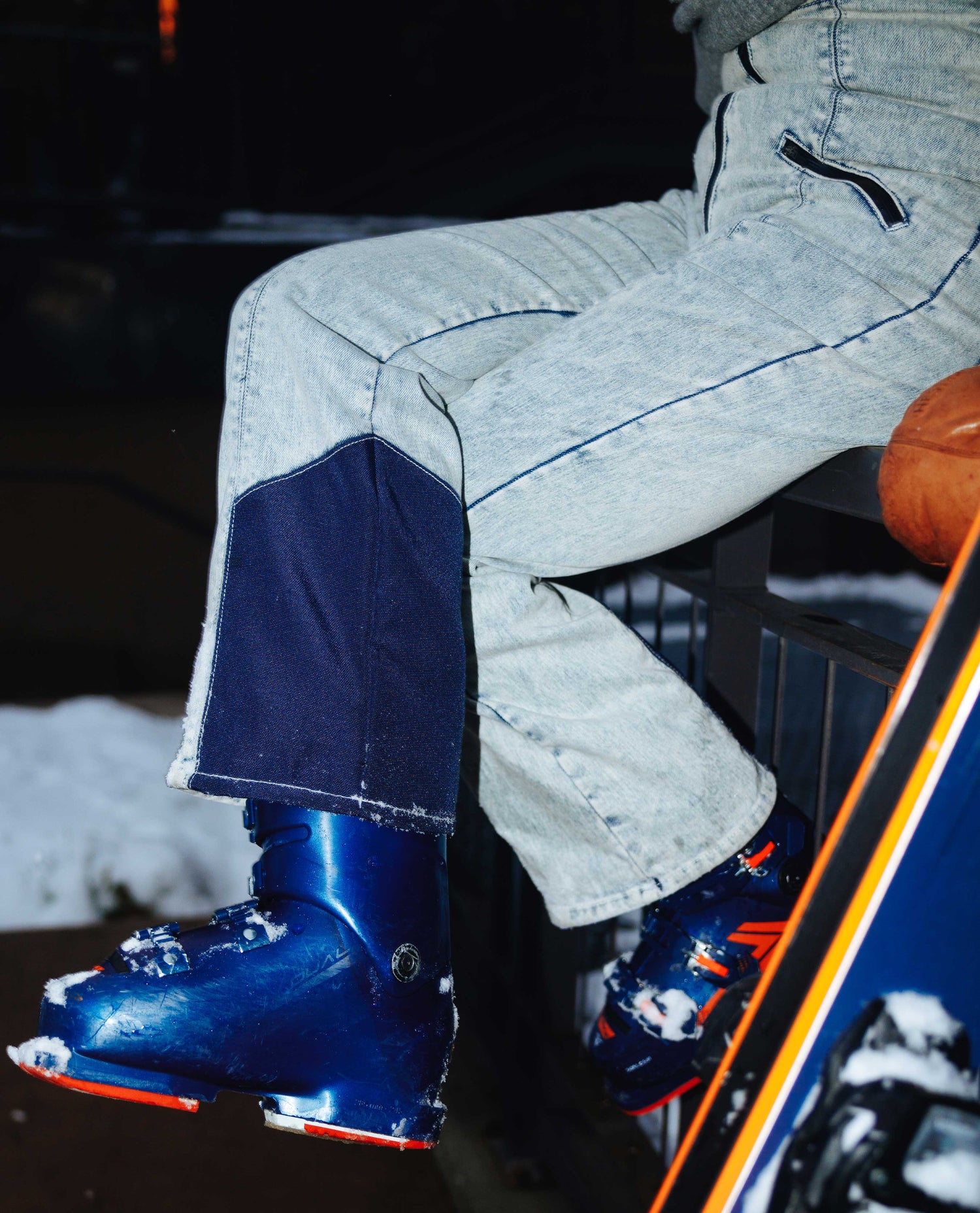 Person wearing light blue jeans and blue ski boots sitting on a snow-covered bench.