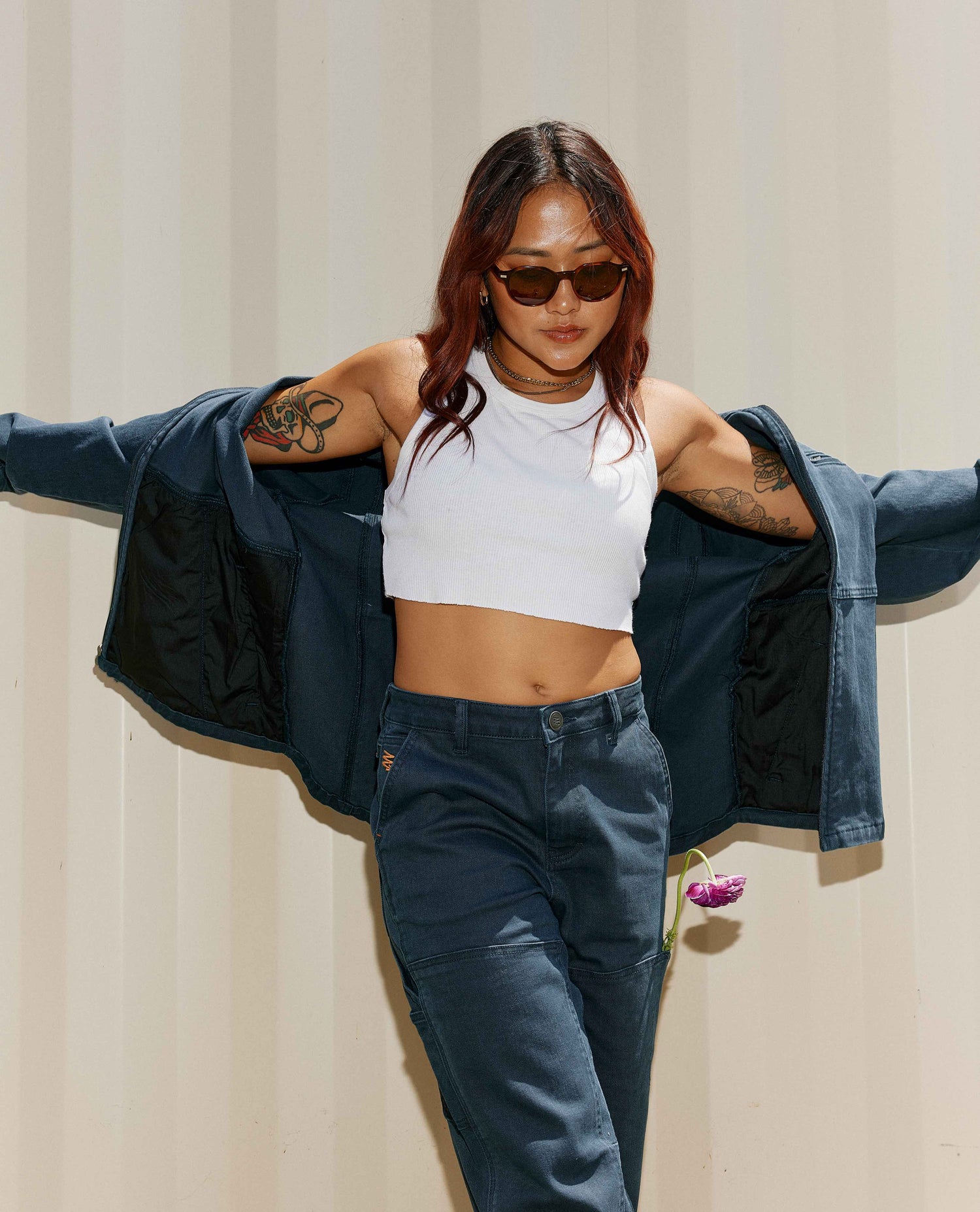A woman in a white crop top, blue cargo style jacket, blue cargo pants, and round brown sunglasses posing with her arms out in front of a tan background with a pink flower in her left pocket. 