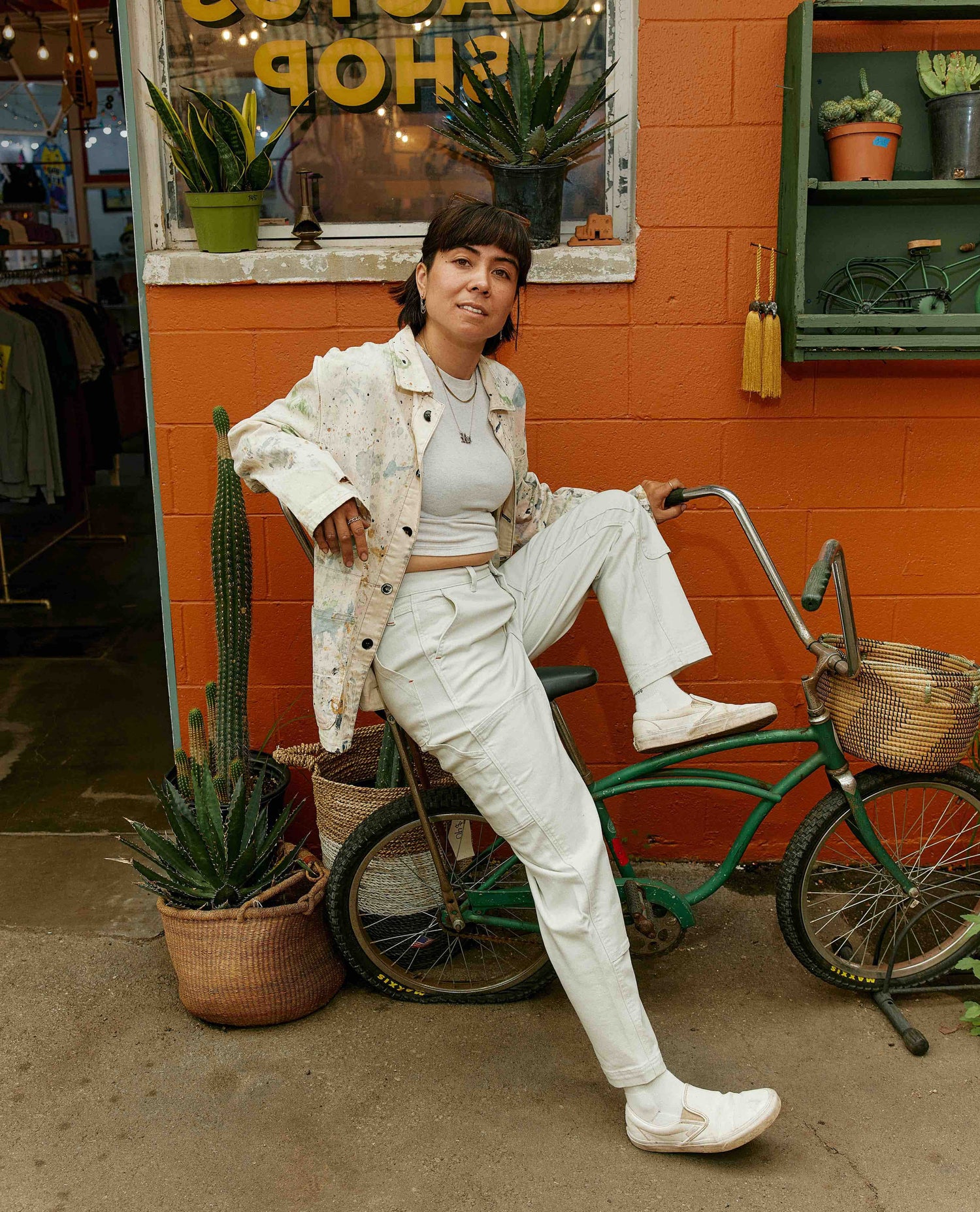 Woman in a white outfit sitting on a green bicycle against an orange wall with plants.