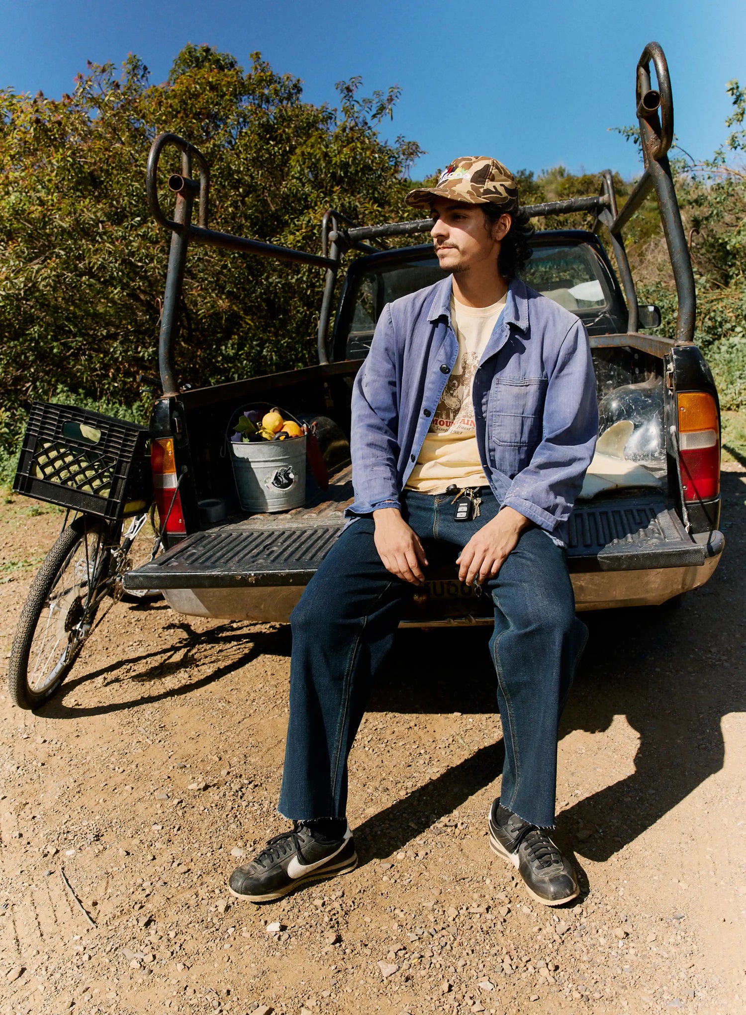 Person sitting in the bed of a pickup truck with a cooler and fruit, surrounded by trees.
