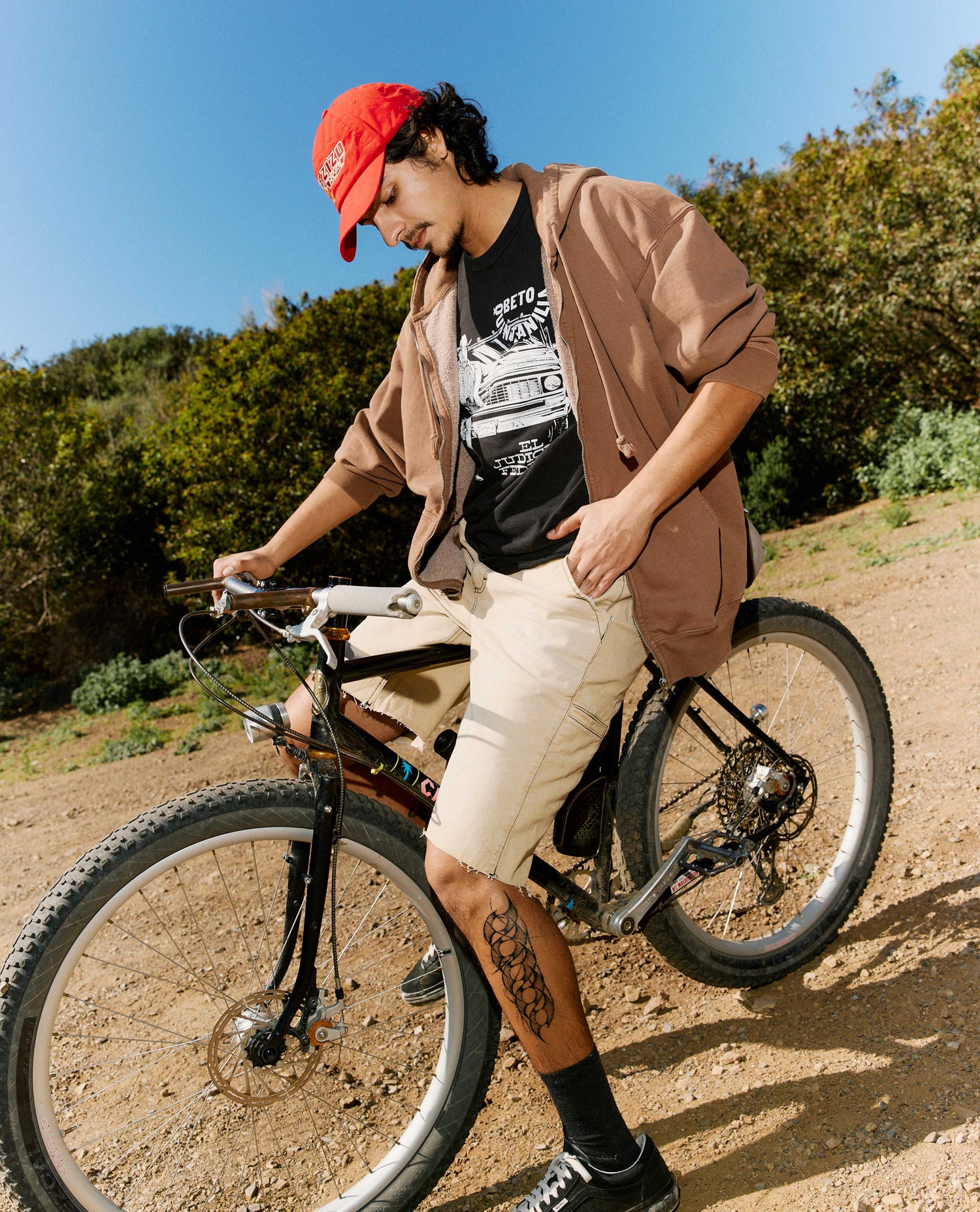 Person with a red cap and tan jacket standing next to a bicycle on a dirt path with trees in the background.