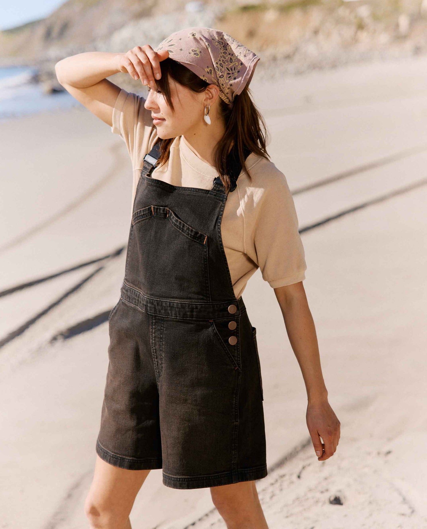Woman wearing a beige shirt, black denim overalls, and a patterned cap on a sandy beach.