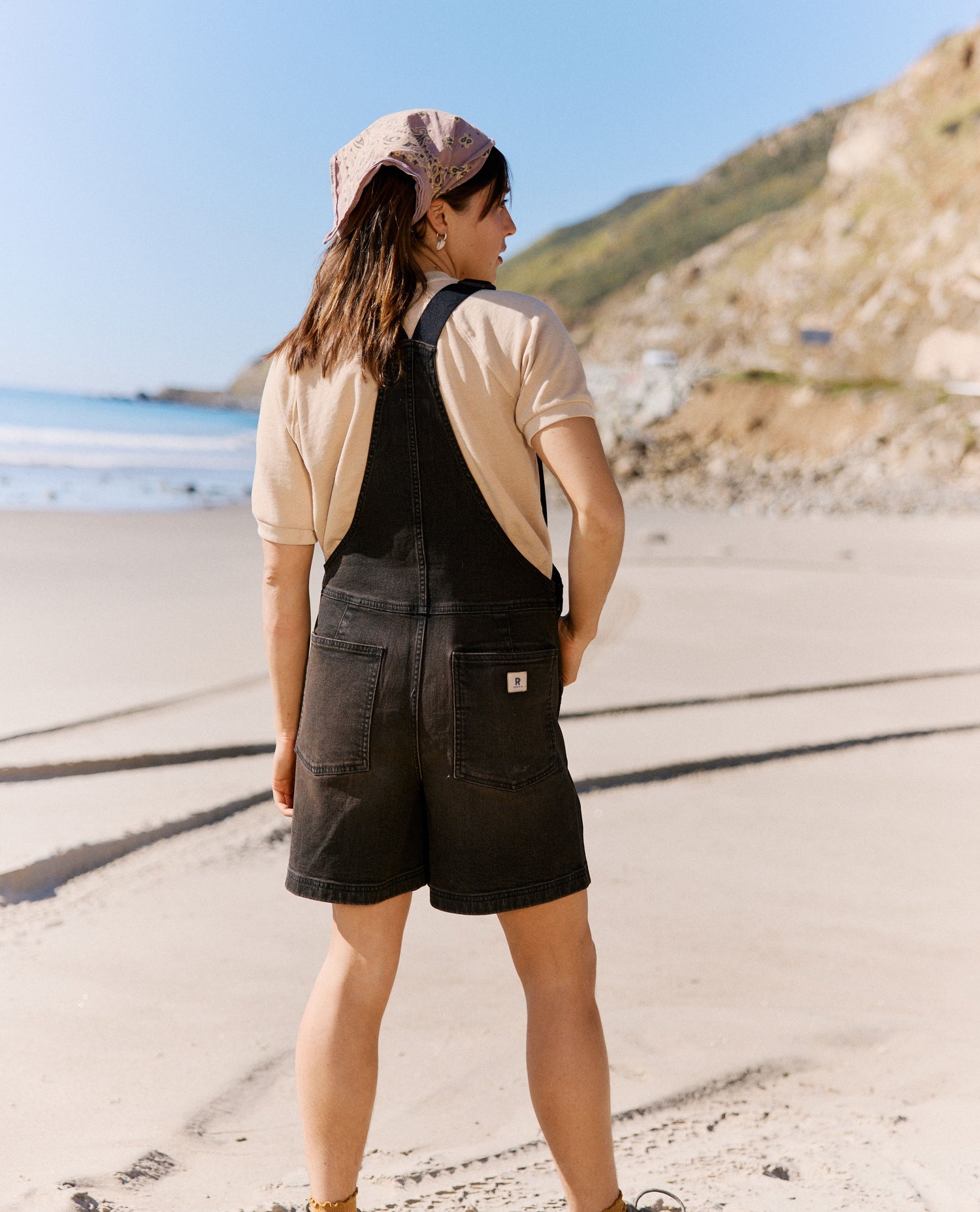 Person wearing black overalls and a cap standing on a beach with mountains in the background