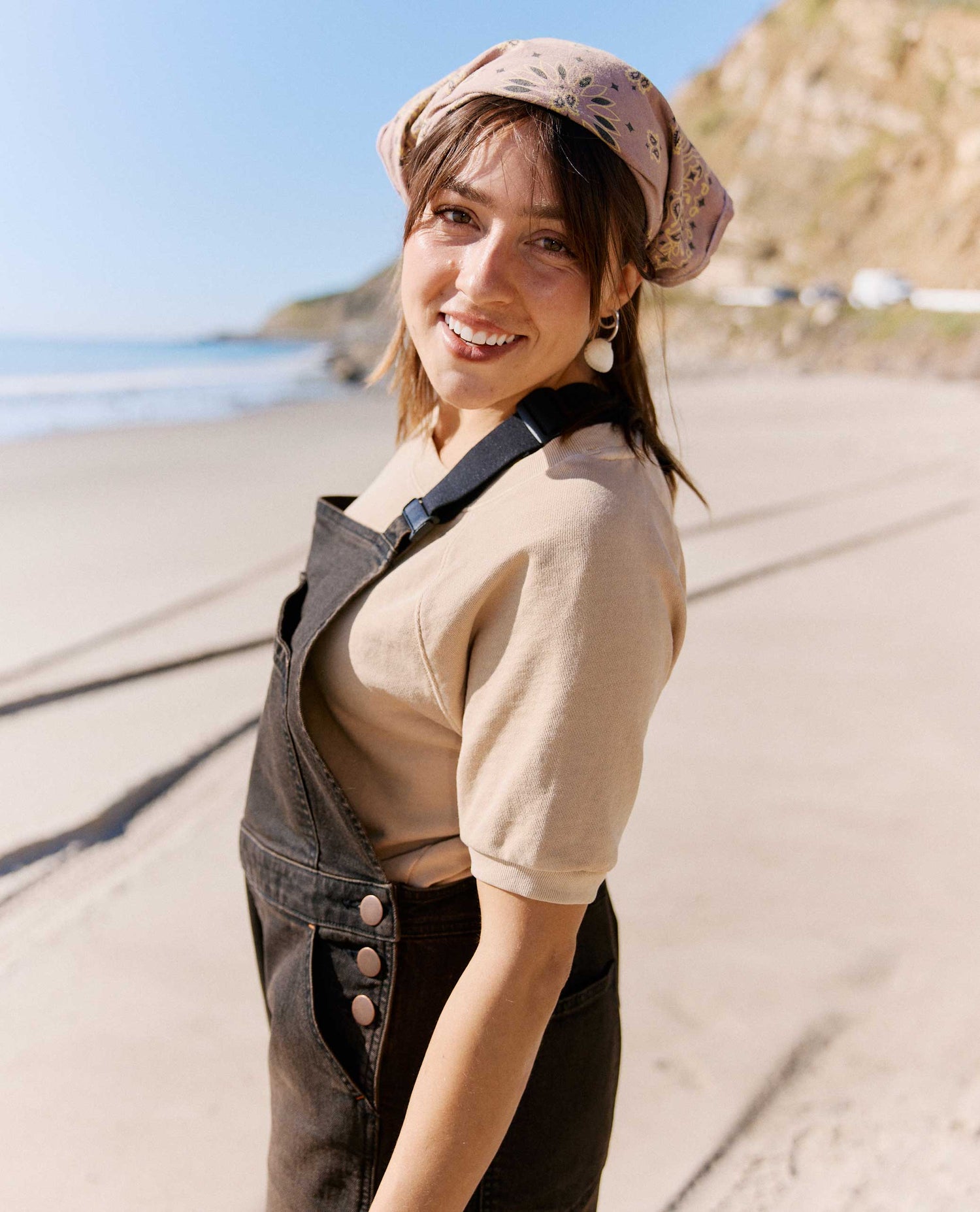 Woman standing on a beach wearing a beige top and denim overalls with a floral headscarf.