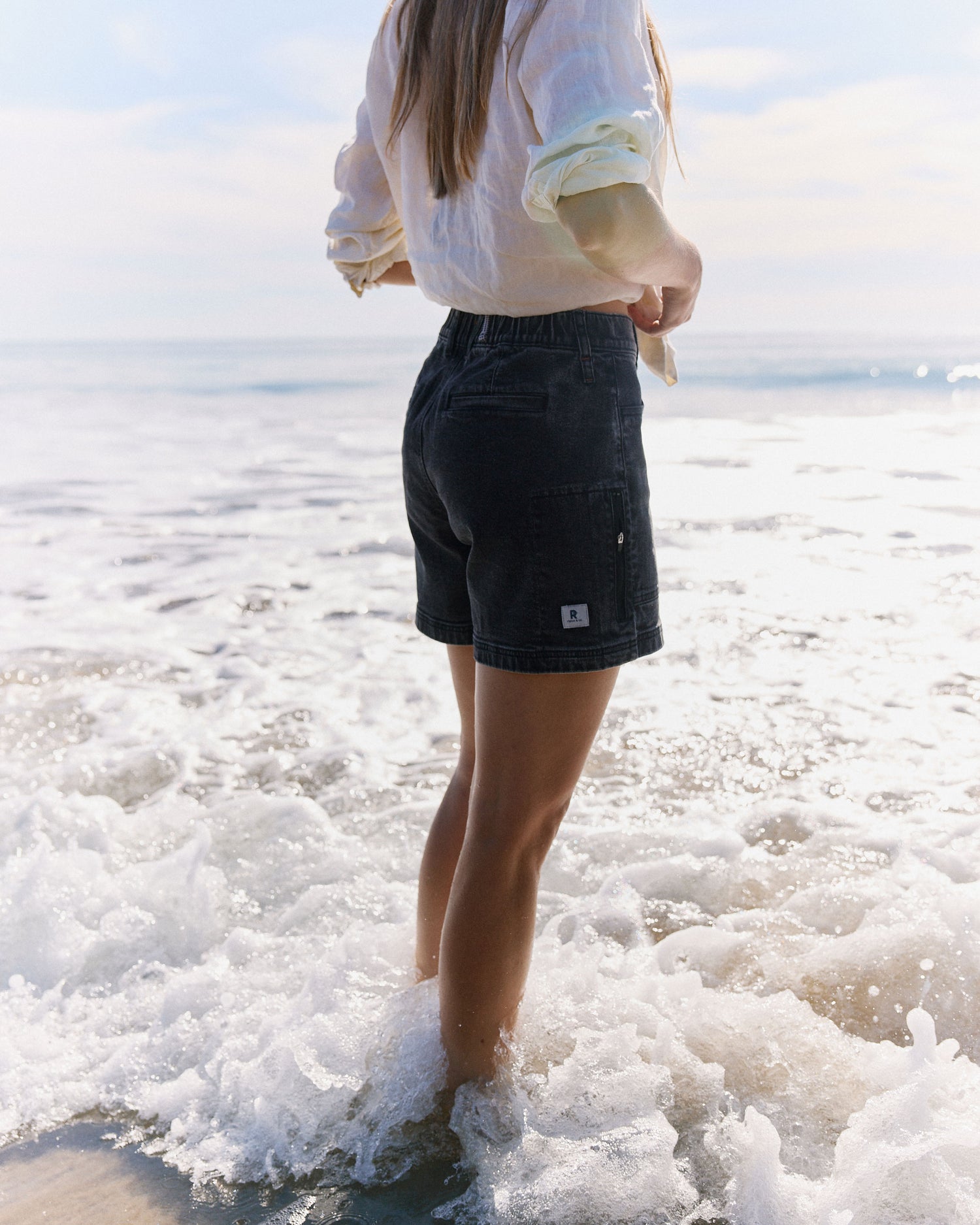 Person wearing a white shirt and black shorts standing in shallow water at the beach.