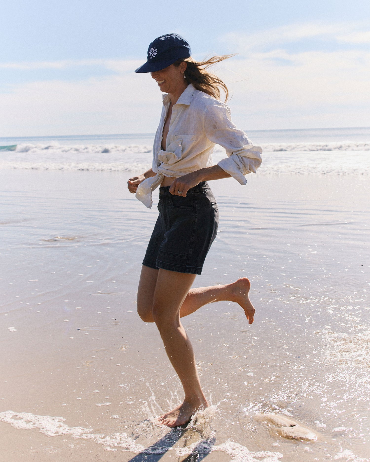 Woman running on a beach wearing a white shirt, black shorts, and a navy cap.