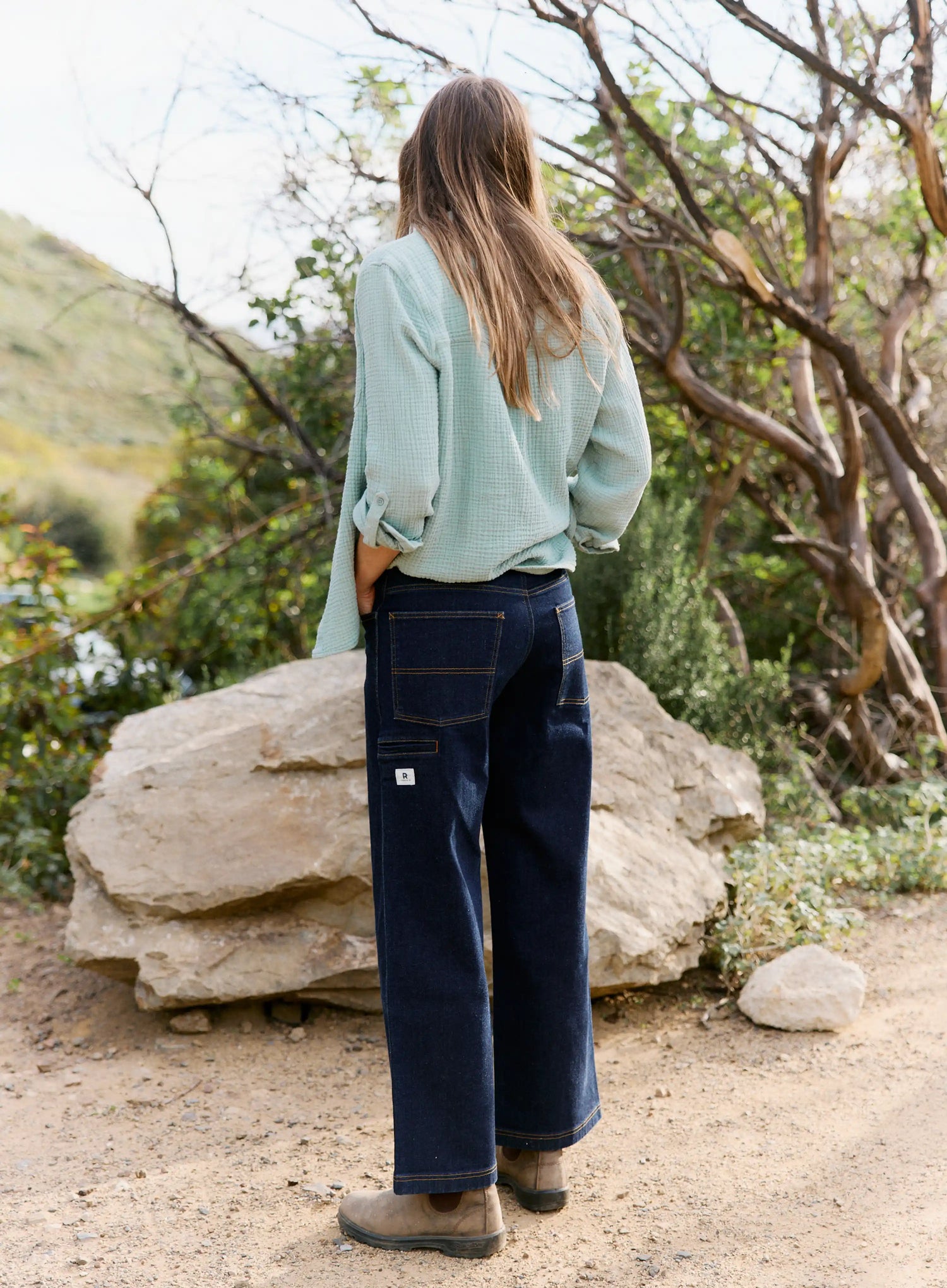 Person wearing a light blue shirt and dark blue jeans standing in a natural setting with trees and rocks.