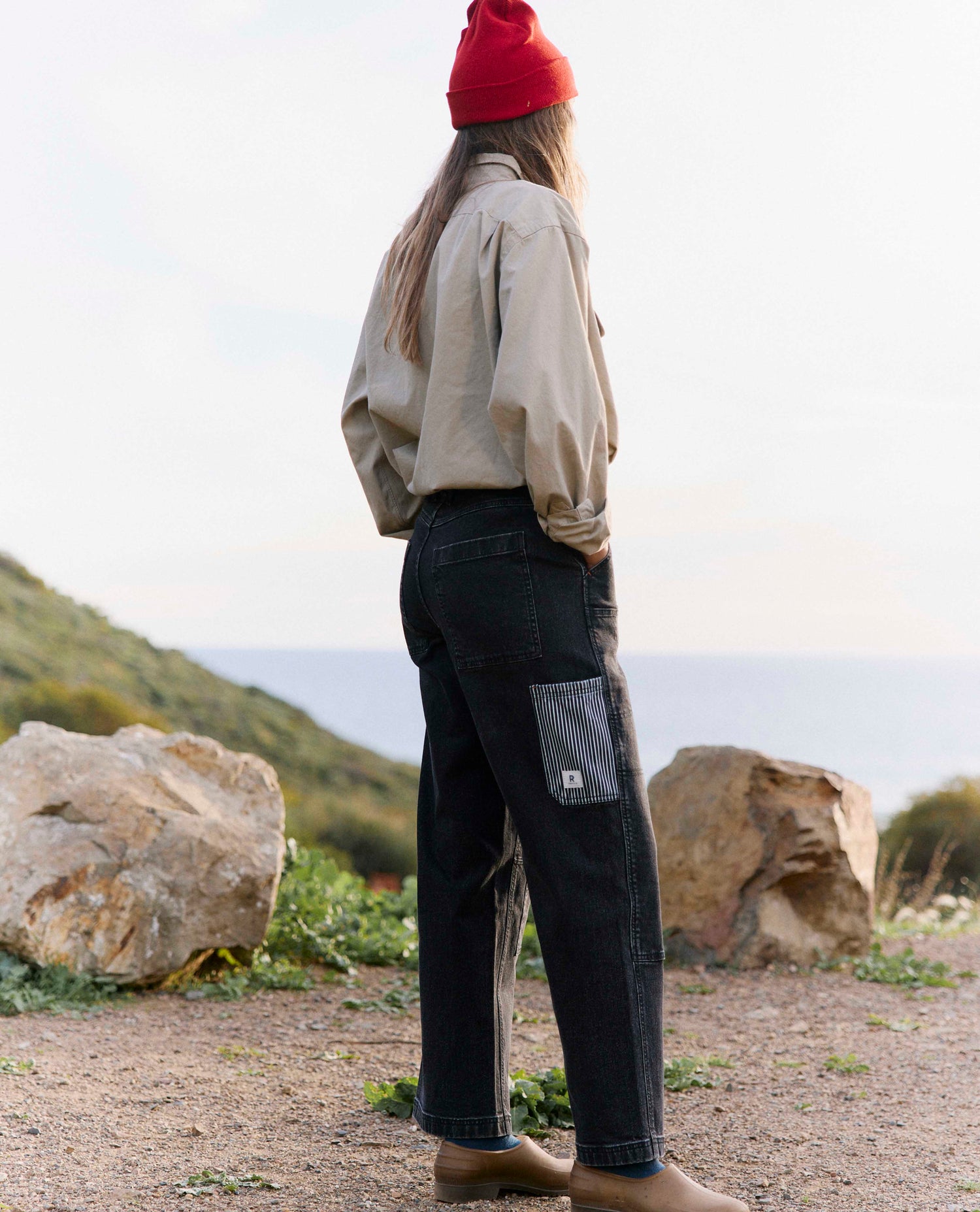 Person wearing a red beanie and beige shirt with dark jeans standing outdoors near rocks and grass.