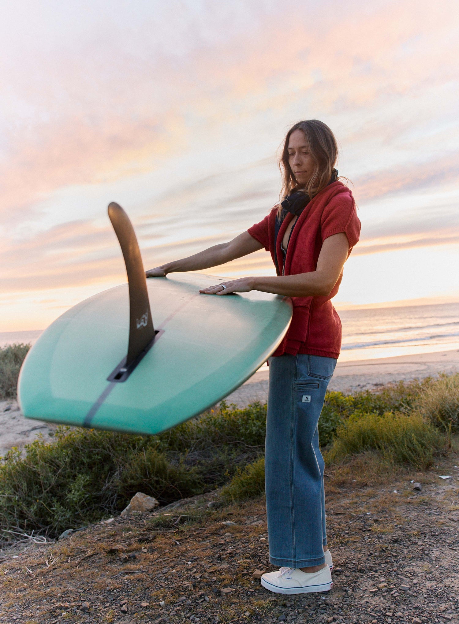 Person holding a teal surfboard on a beach at sunset