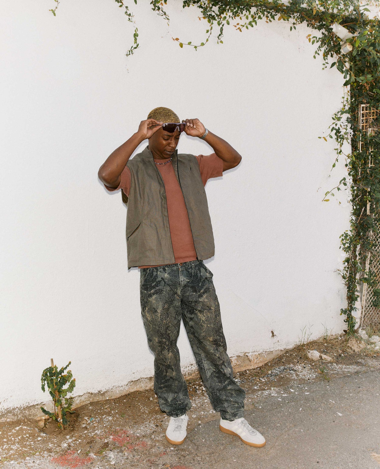 Person wearing a brown shirt, green vest, and camouflage pants against a white wall with greenery.