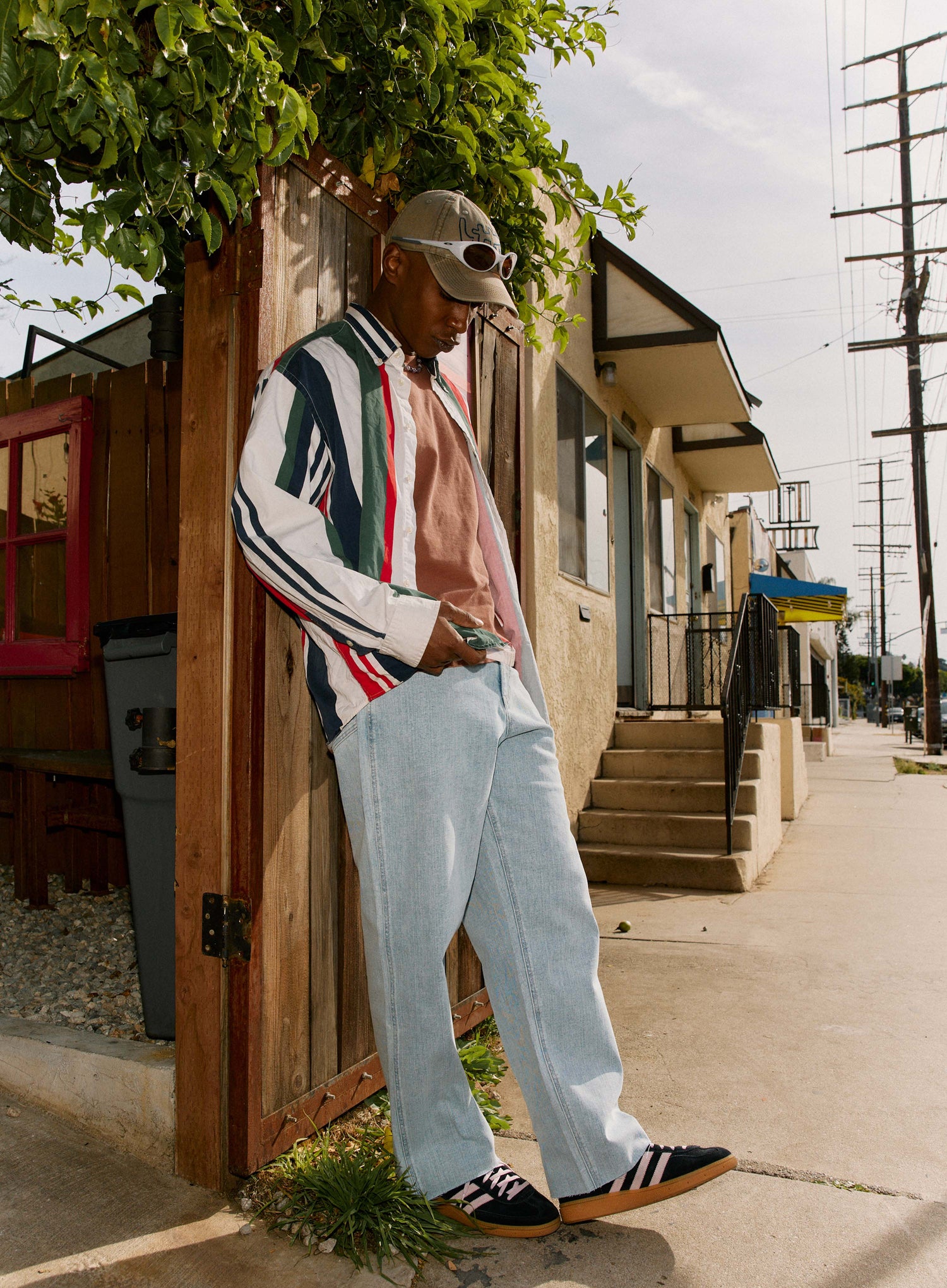 Person wearing a colorful jacket and light blue pants standing on a sidewalk with a wooden door and building in the background.
