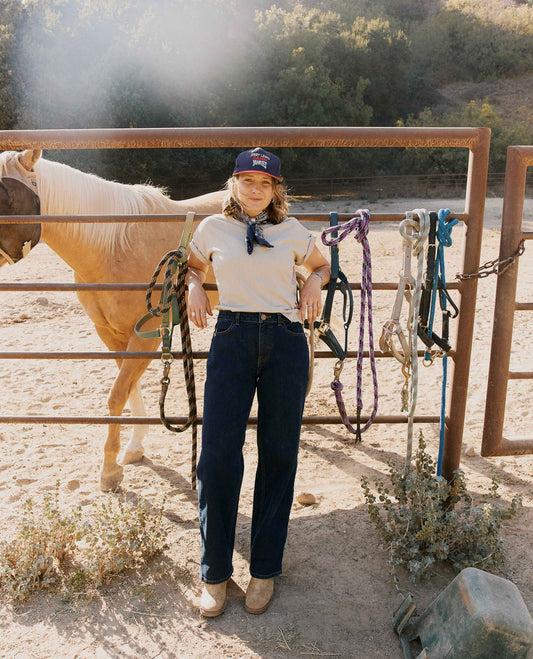 A woman stands in a dusty outdoor ranch setting next to a horse, wearing a white t-shirt, dark blue jeans, and a navy cap. She is holding ropes hanging from a metal gate, with a horse and trees in the background.