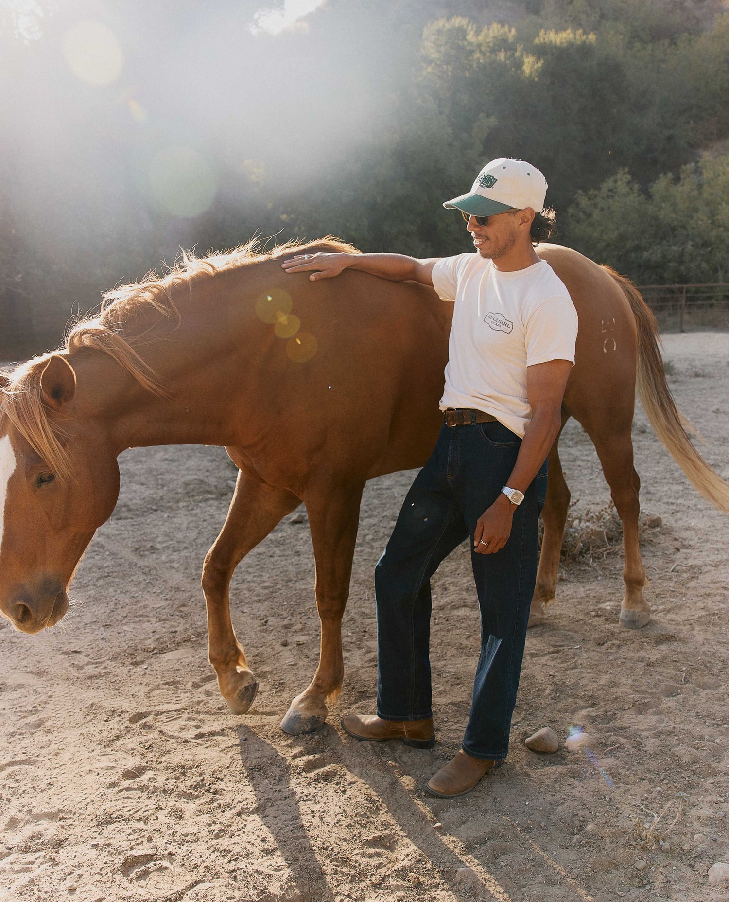 A man wearing a white t-shirt, dark blue jeans, and a baseball cap stands next to a brown horse in a sandy outdoor paddock during golden hour.