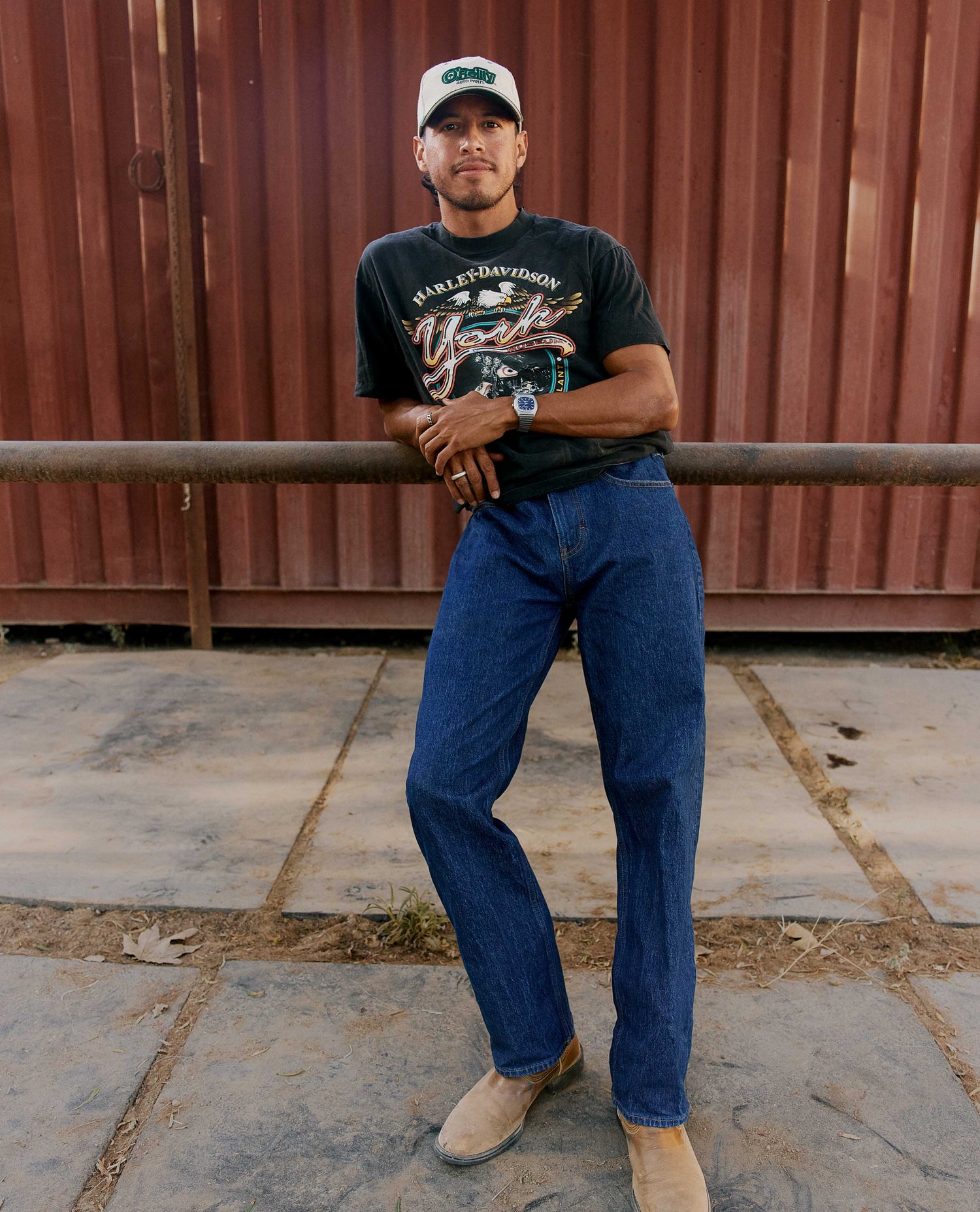 A man wearing a black graphic t-shirt, blue jeans, and tan boots stands outdoors against a corrugated metal wall, leaning on a horizontal bar.