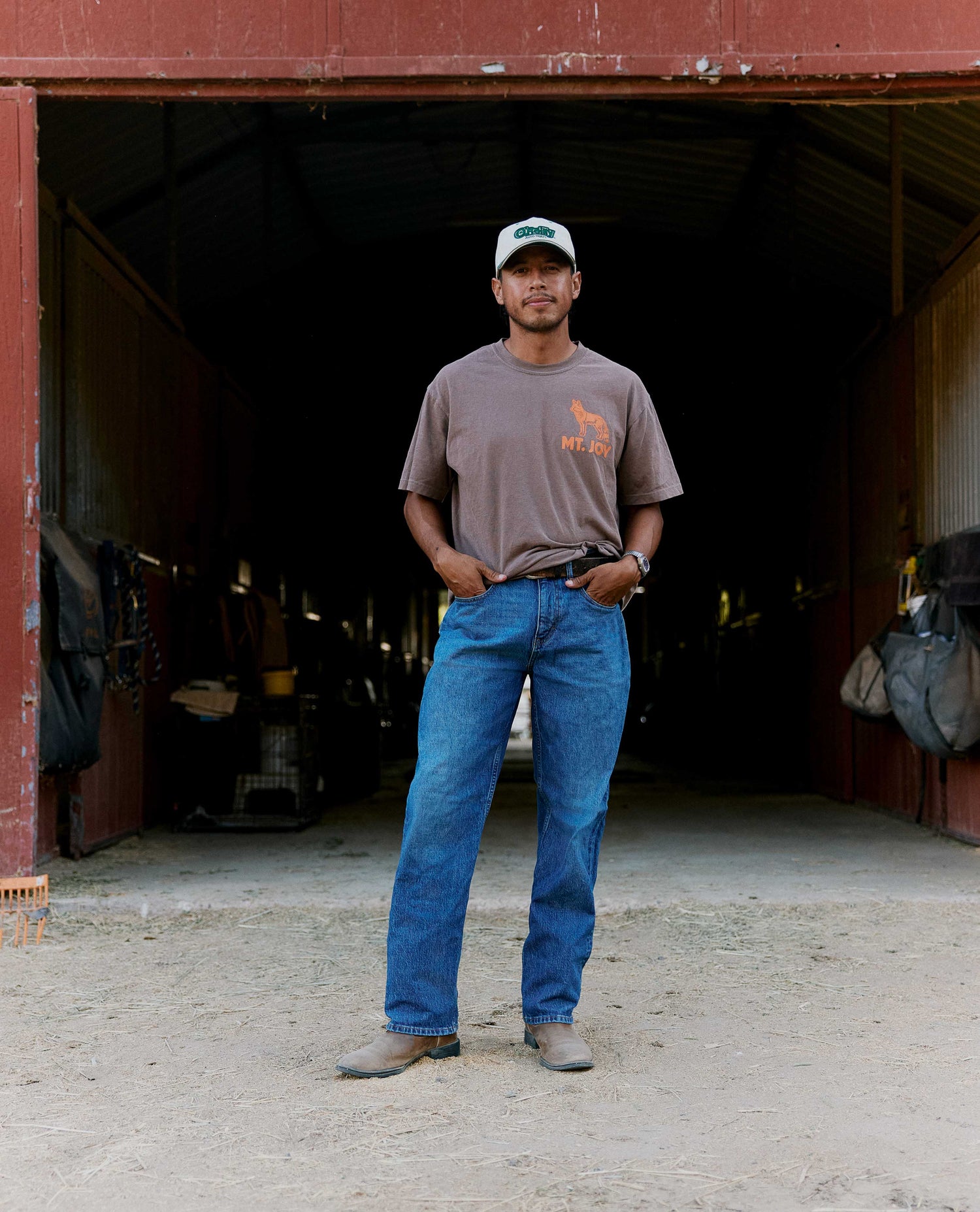 A man stands in the doorway of a rustic barn wearing a brown t-shirt with an orange graphic, blue jeans, and a white cap.