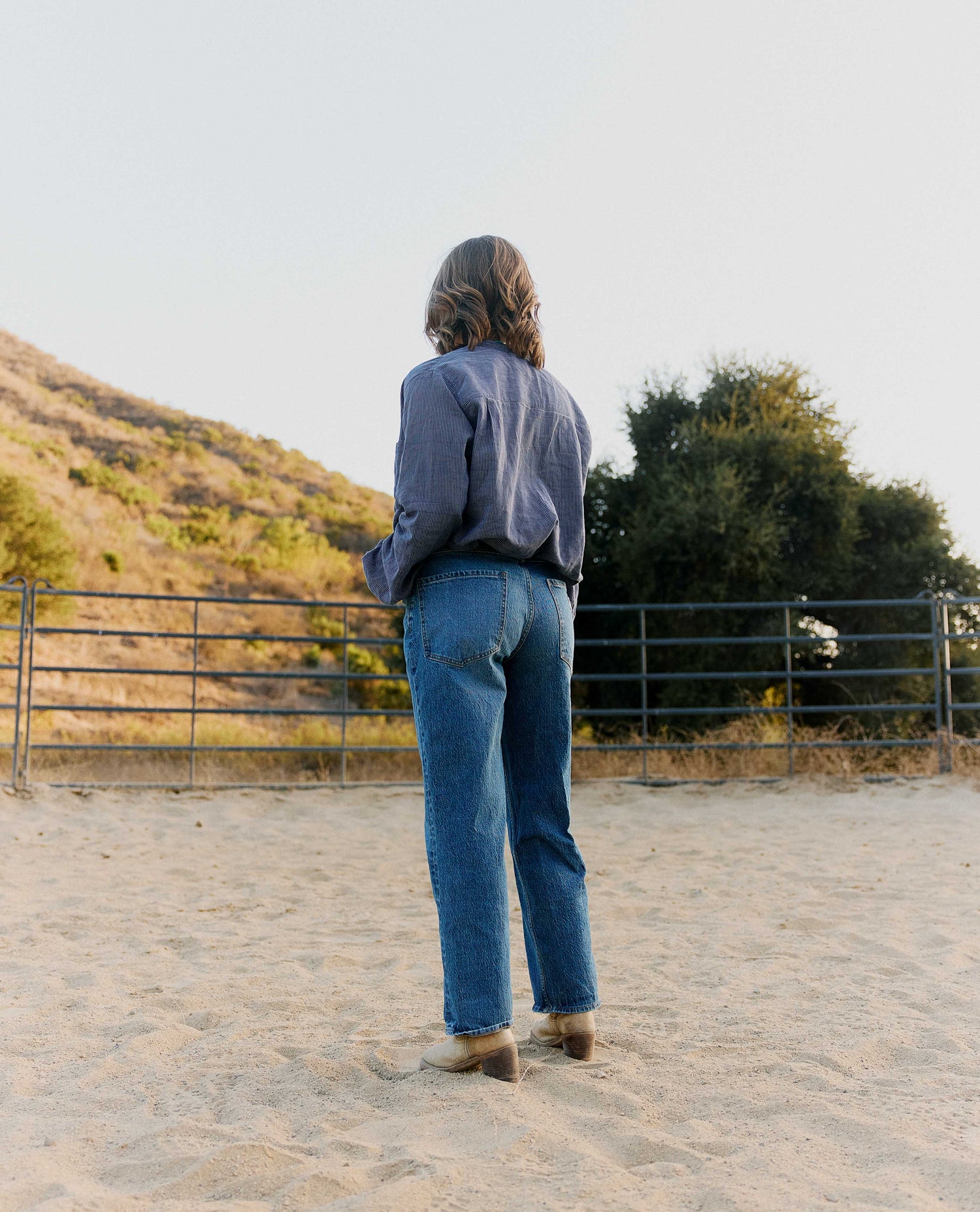 A person standing on sand facing a fence and hillside, wearing a blue button-up shirt and blue jeans.