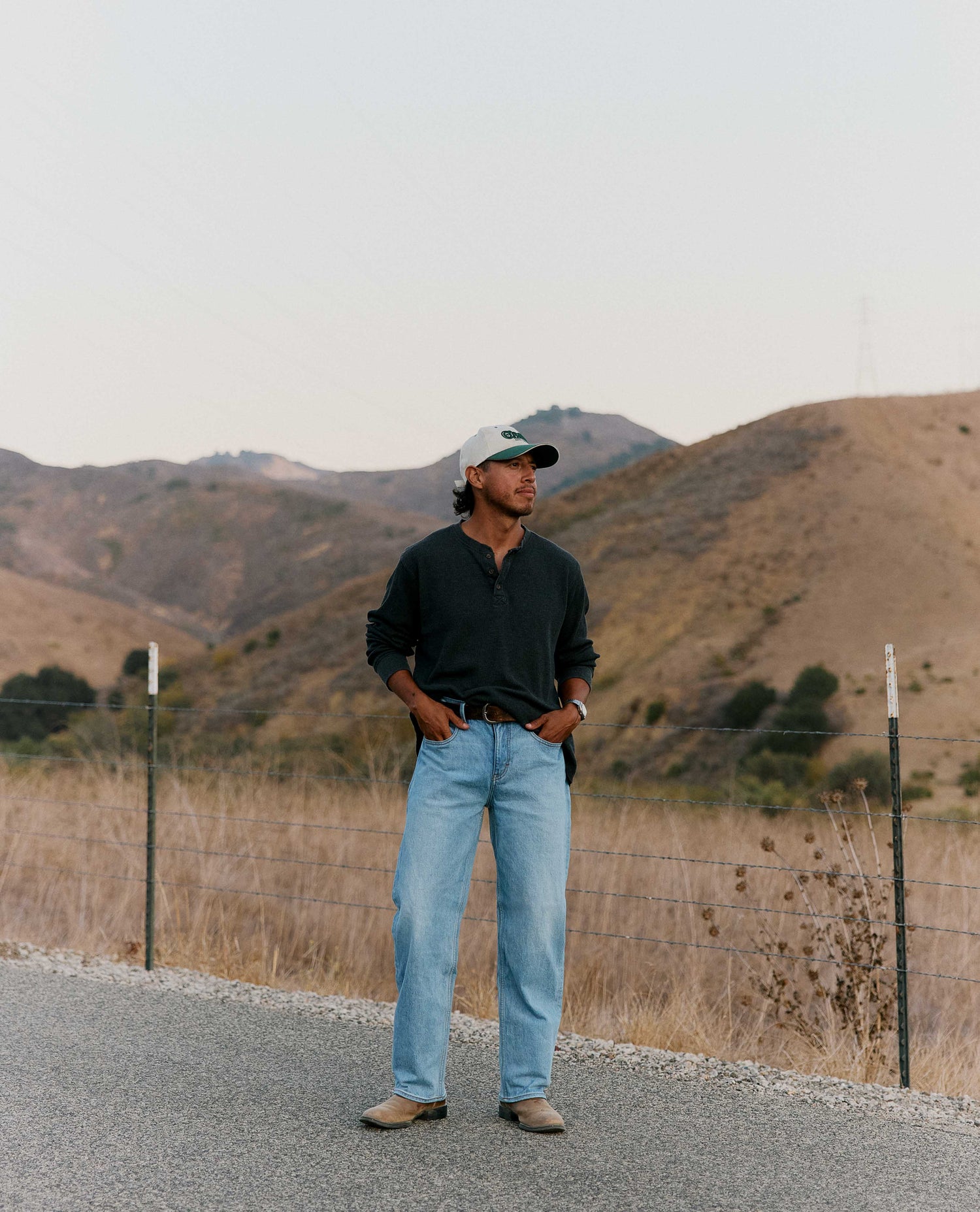 A man stands on a gravel road in a rural landscape with dry hills in the background, wearing a dark long-sleeve henley shirt, light blue jeans, and a baseball cap.