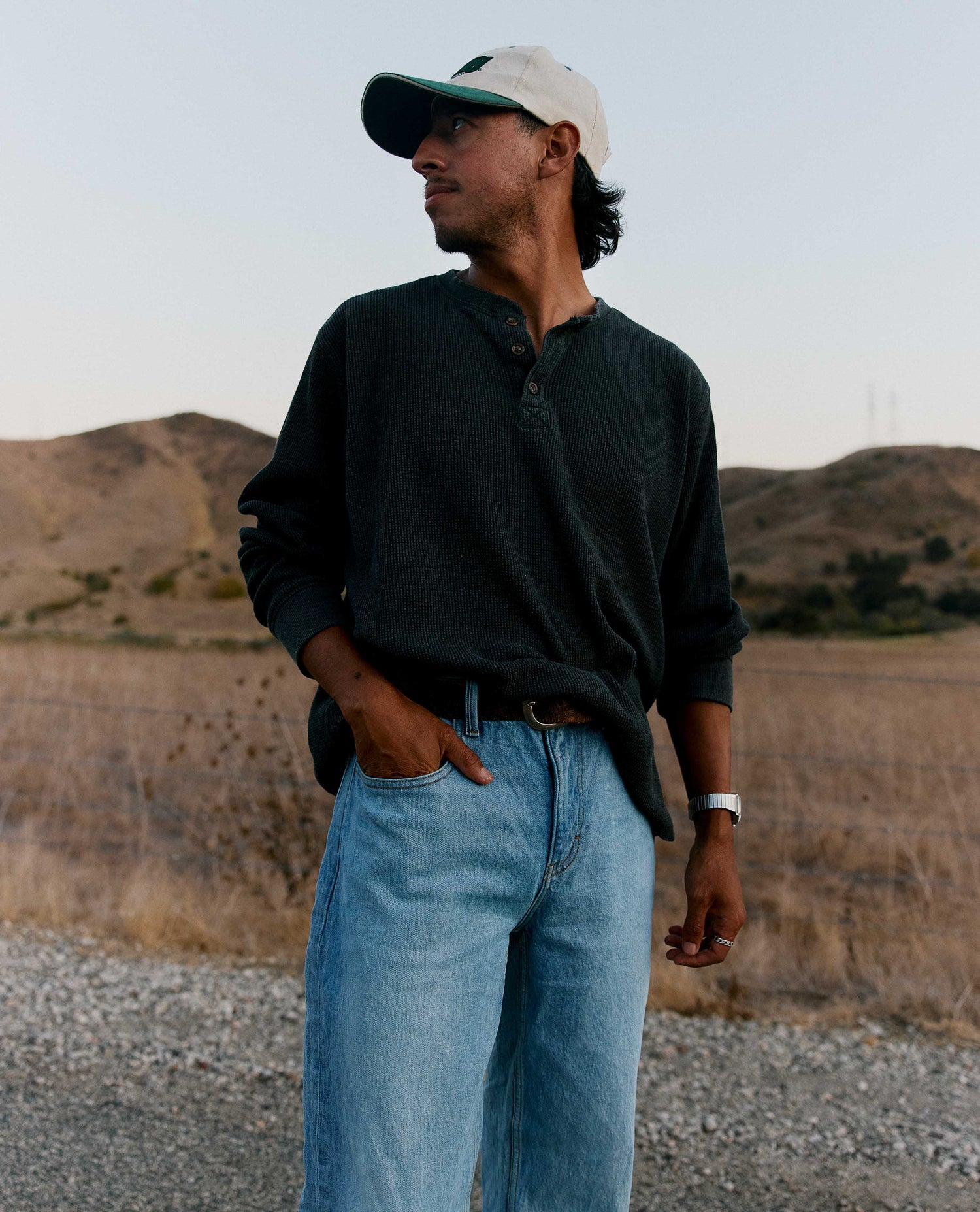 A man wearing a dark charcoal henley shirt, light blue jeans, and a beige baseball cap stands outdoors in a dry, hilly landscape.