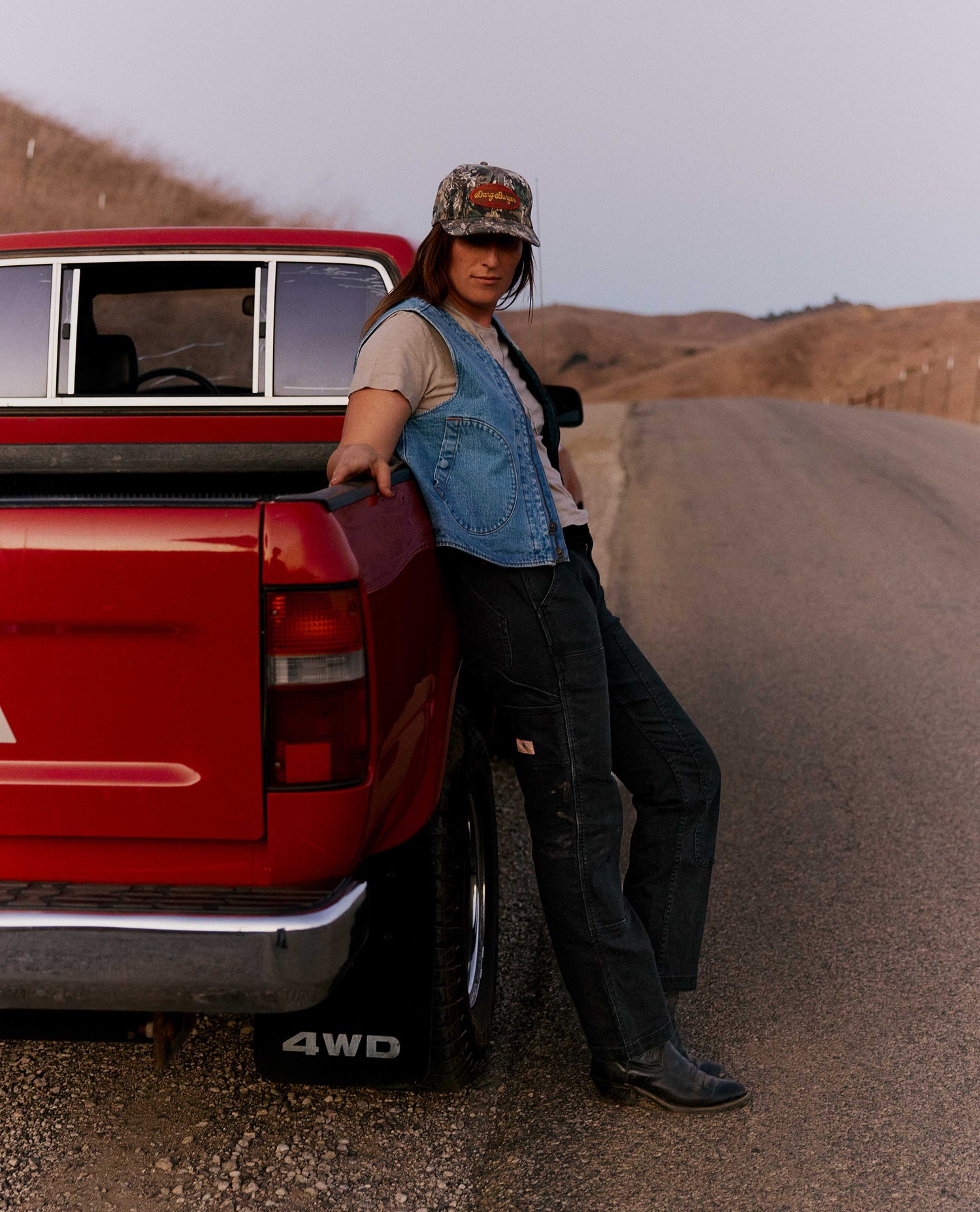 Woman leaning against a red pickup truck wearing a blue denim vest and a camo hat