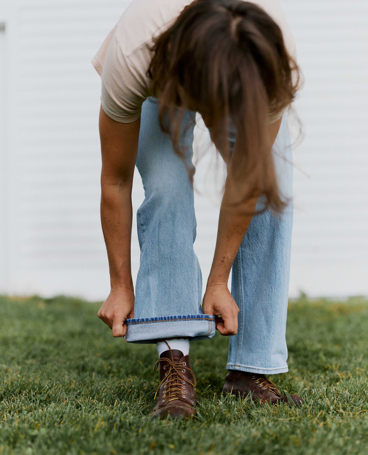 Woman standing in front of white barn in green grass, wearing light wash blue jeans and folding her jean cuffs. She has brown boots and a tan shirt on. 