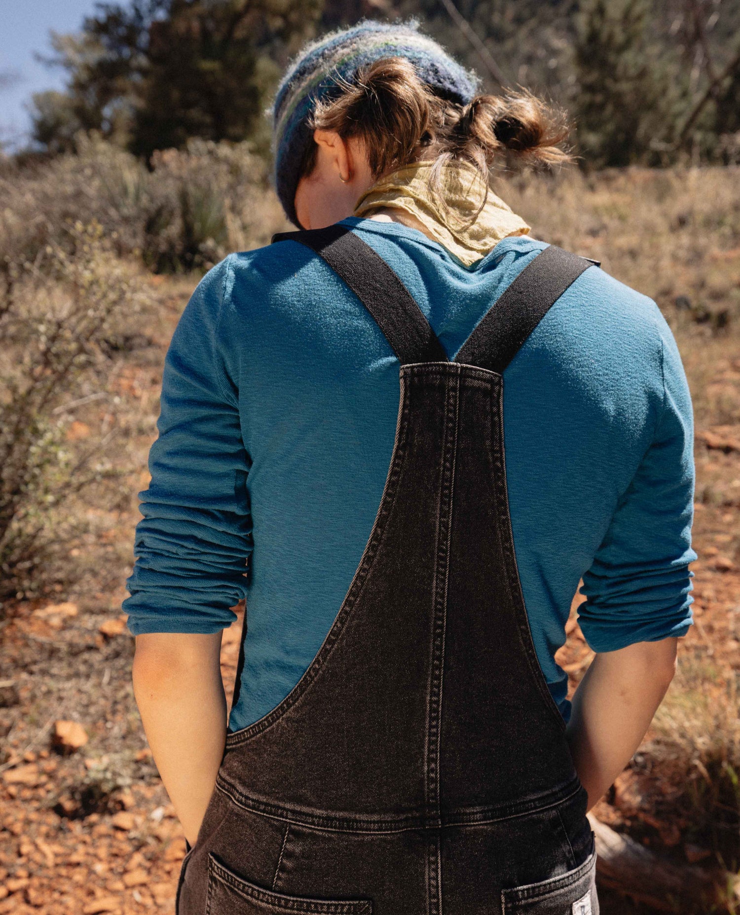 Person wearing a blue shirt and black overalls standing in a desert-like landscape.