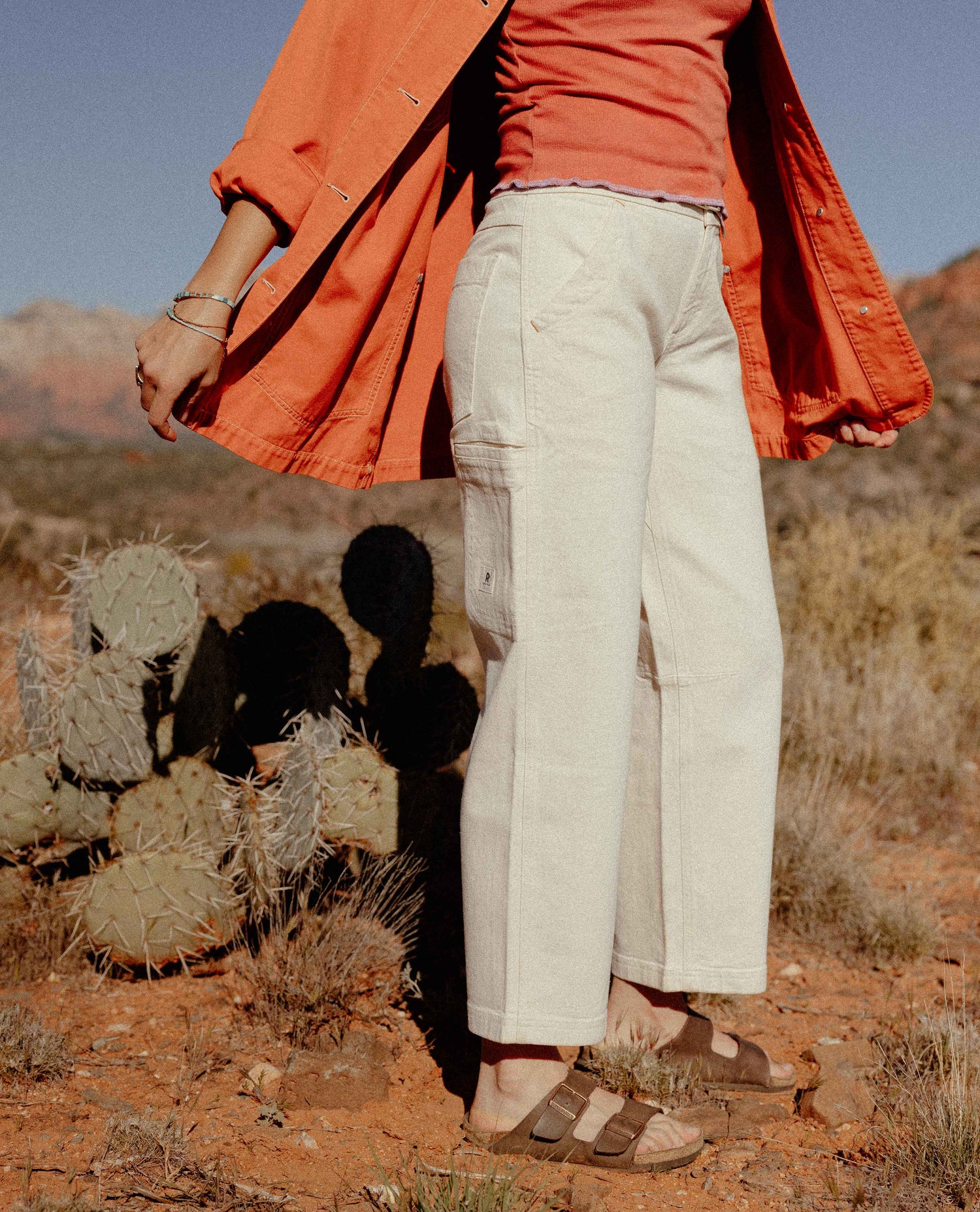 Person wearing an orange coat and white pants standing in a desert landscape with cacti.