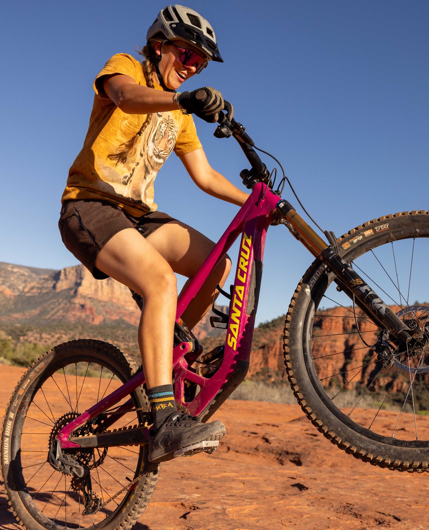 Person riding a pink Santa Cruz mountain bike on a dirt trail with red rock formations in the background.