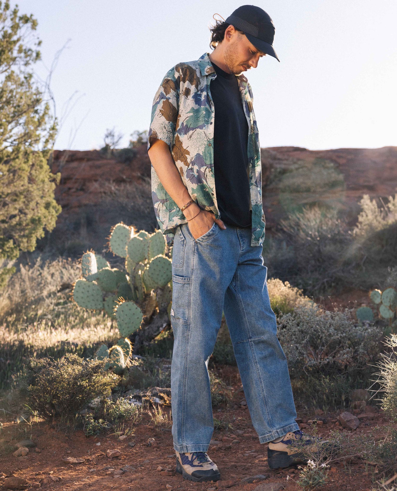 Man outside in desert wearing black shirt and hat, blue jeans, and multicolored shirt. 