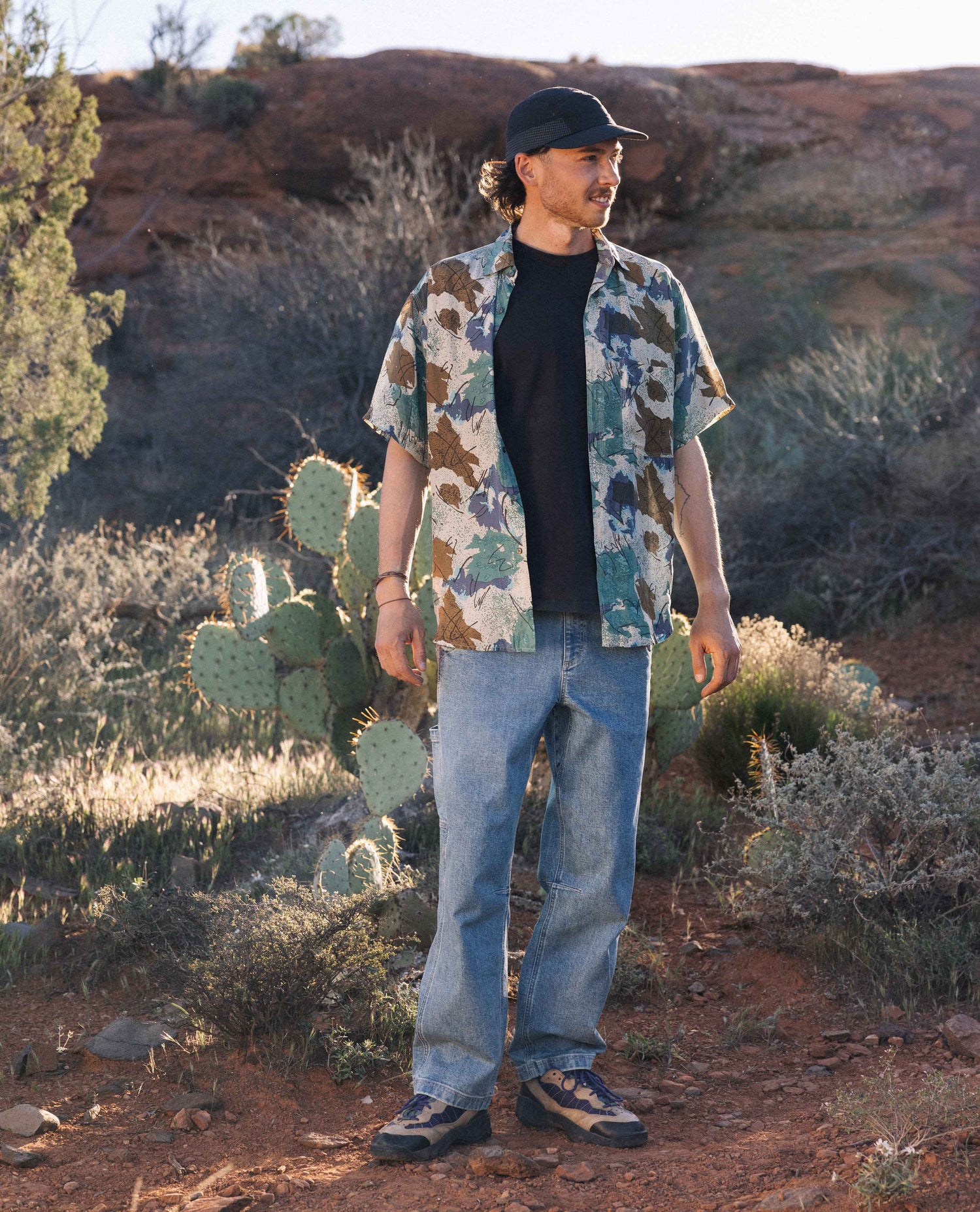 Man standing in desert, in front of cactus, wearing black hat and shirt, blue jeans, and multicolored shirt. 