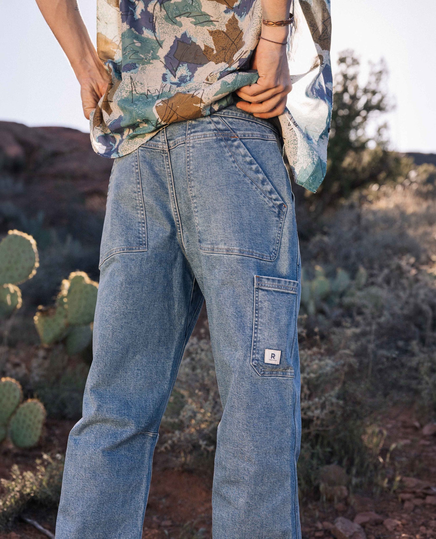 Man outside in front of cactus, with hands on waistband of a pair of blue jeans.