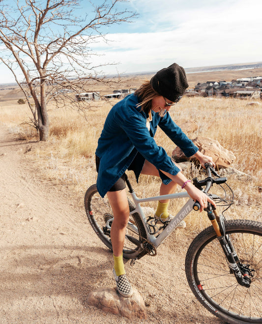 Girl on bike with blue shirt, black shorts, outside on a gravel trail with tree in background