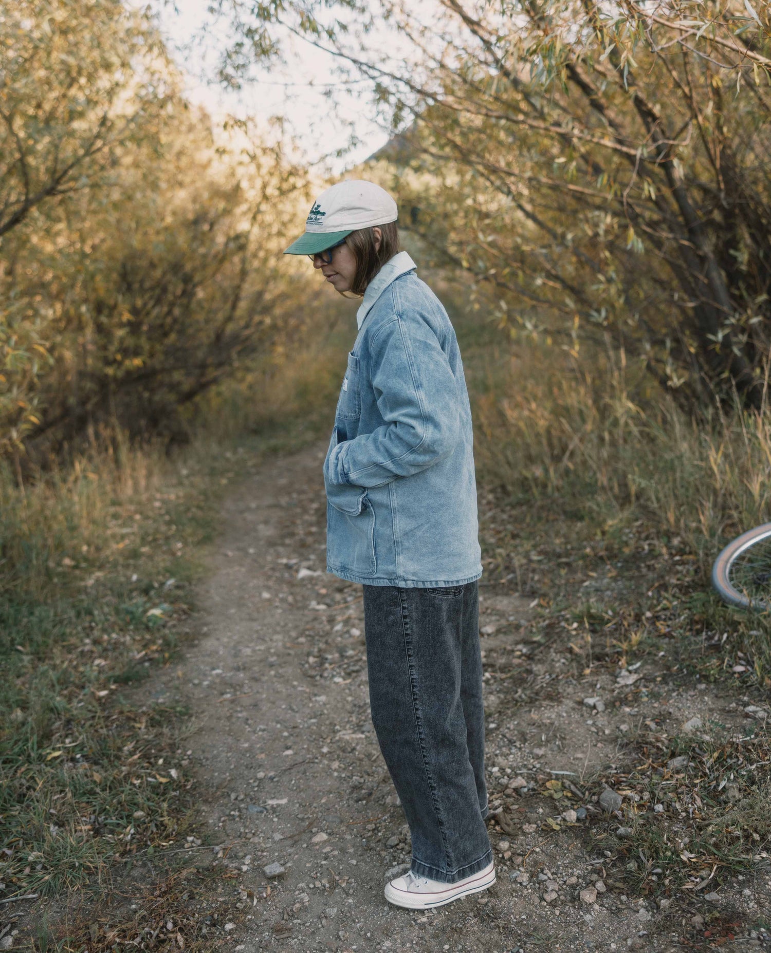 Person standing on a dirt path in a natural setting wearing a denim jacket and cap. Color:Light