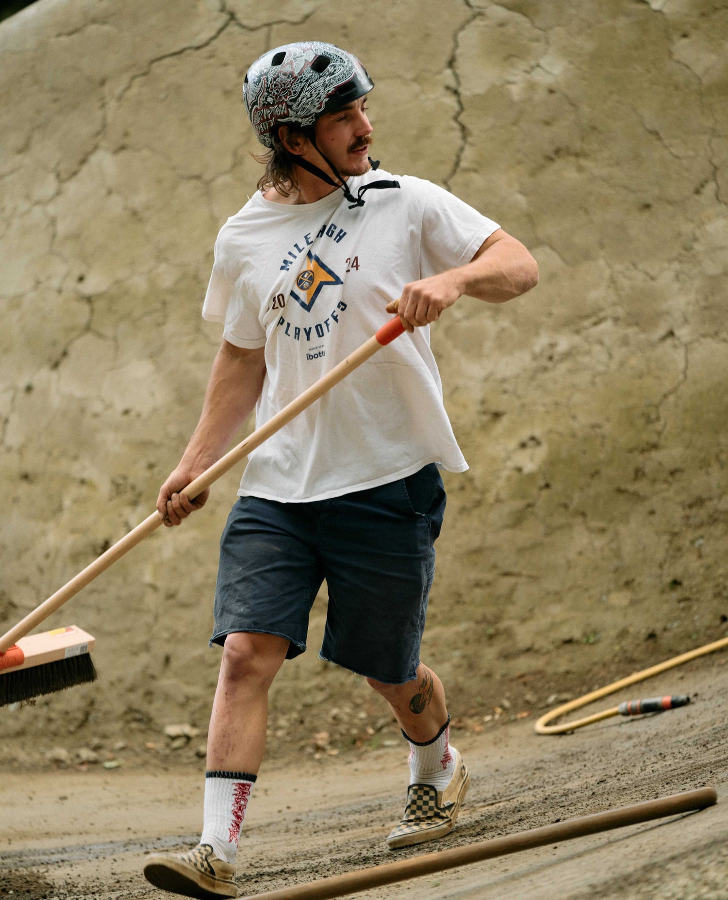 Person holding a broom in a dirt area with a helmet on