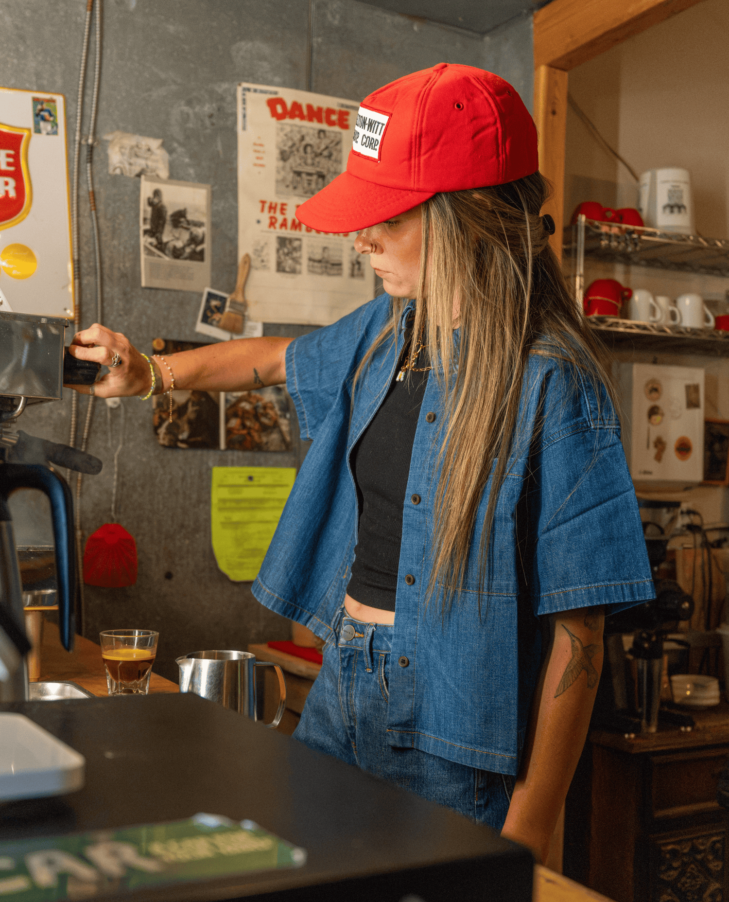 A woman wearing a red baseball cap, a blue denim shirt over a black crop top, and blue jeans stands behind a counter in a cafe, pouring coffee from a machine.