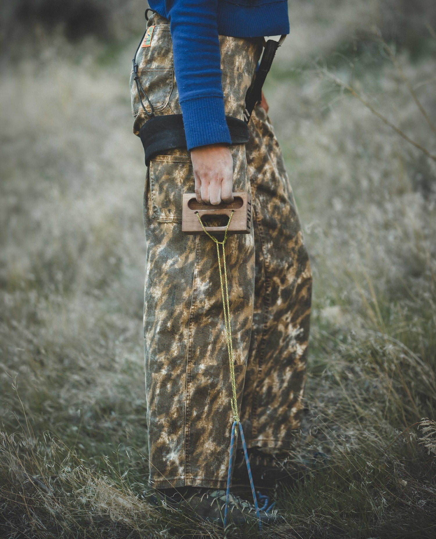 Person holding a bow in camouflage pants and blue sweater outdoors