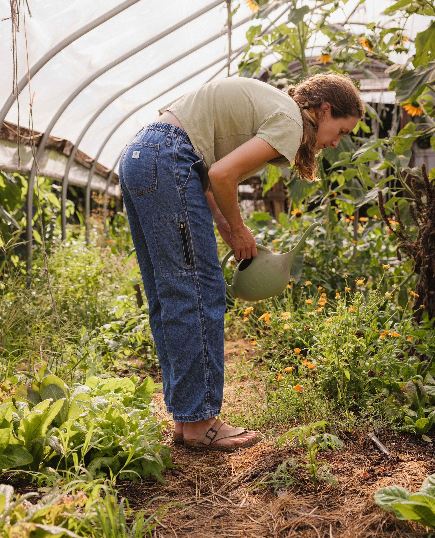 A woman watering plants in a greenhouse wearing blue denim cargo pants, a light olive green t-shirt, and brown sandals.