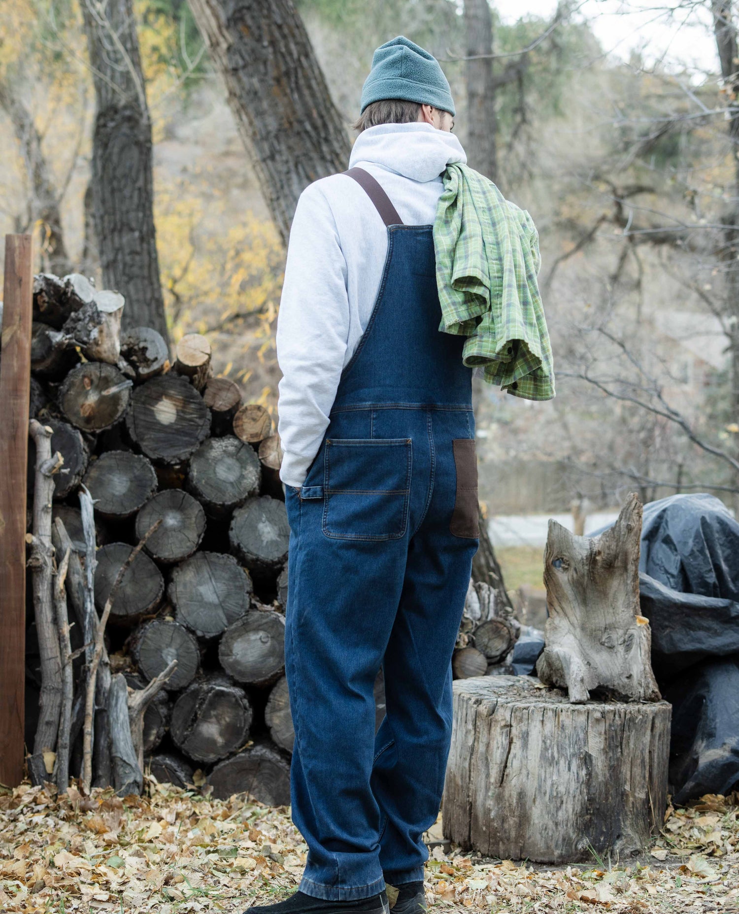 Person wearing blue overalls and a green beanie standing near stacked logs in a forest setting.