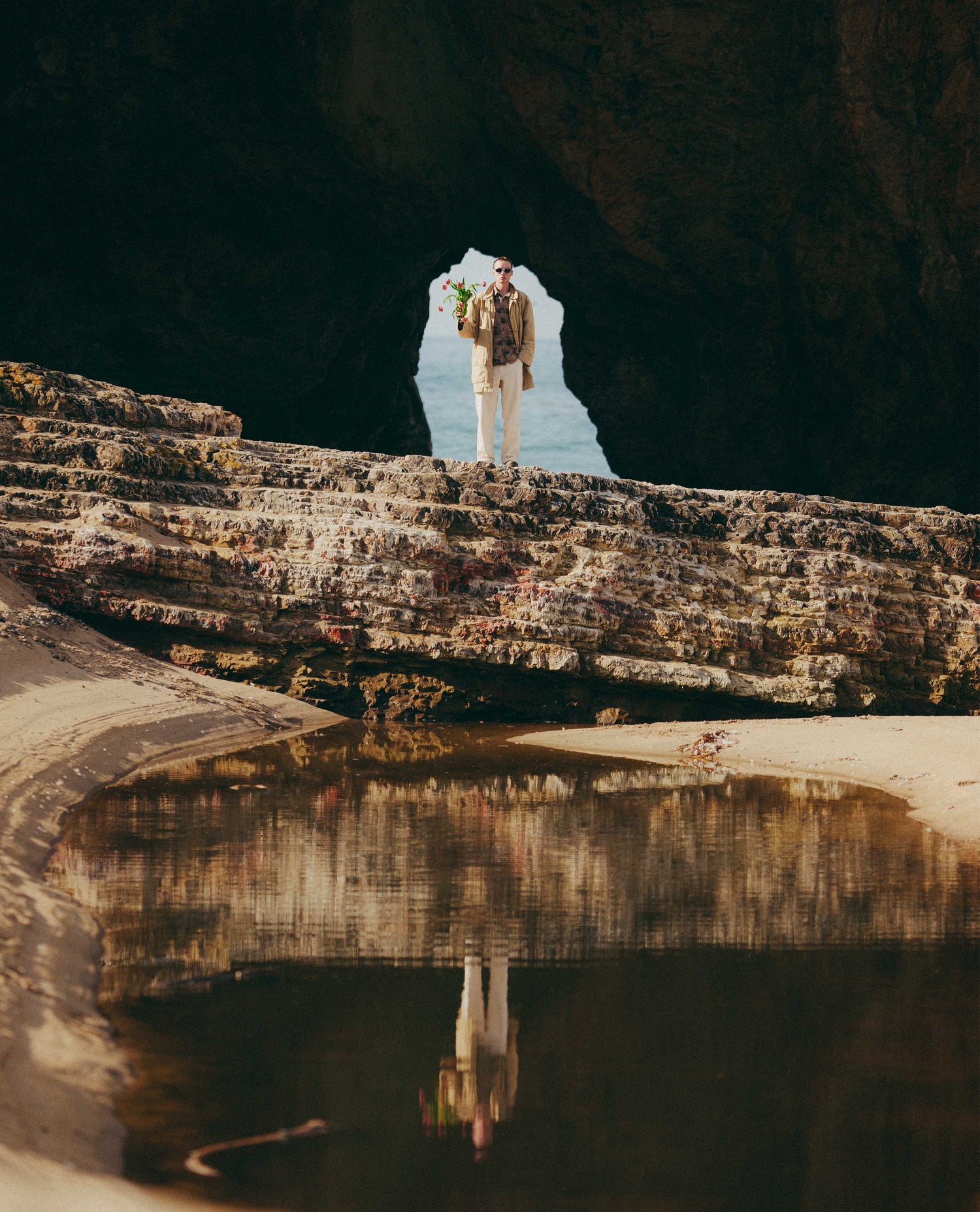 Person standing on a rocky outcrop with a cave in the background, reflected in water below.