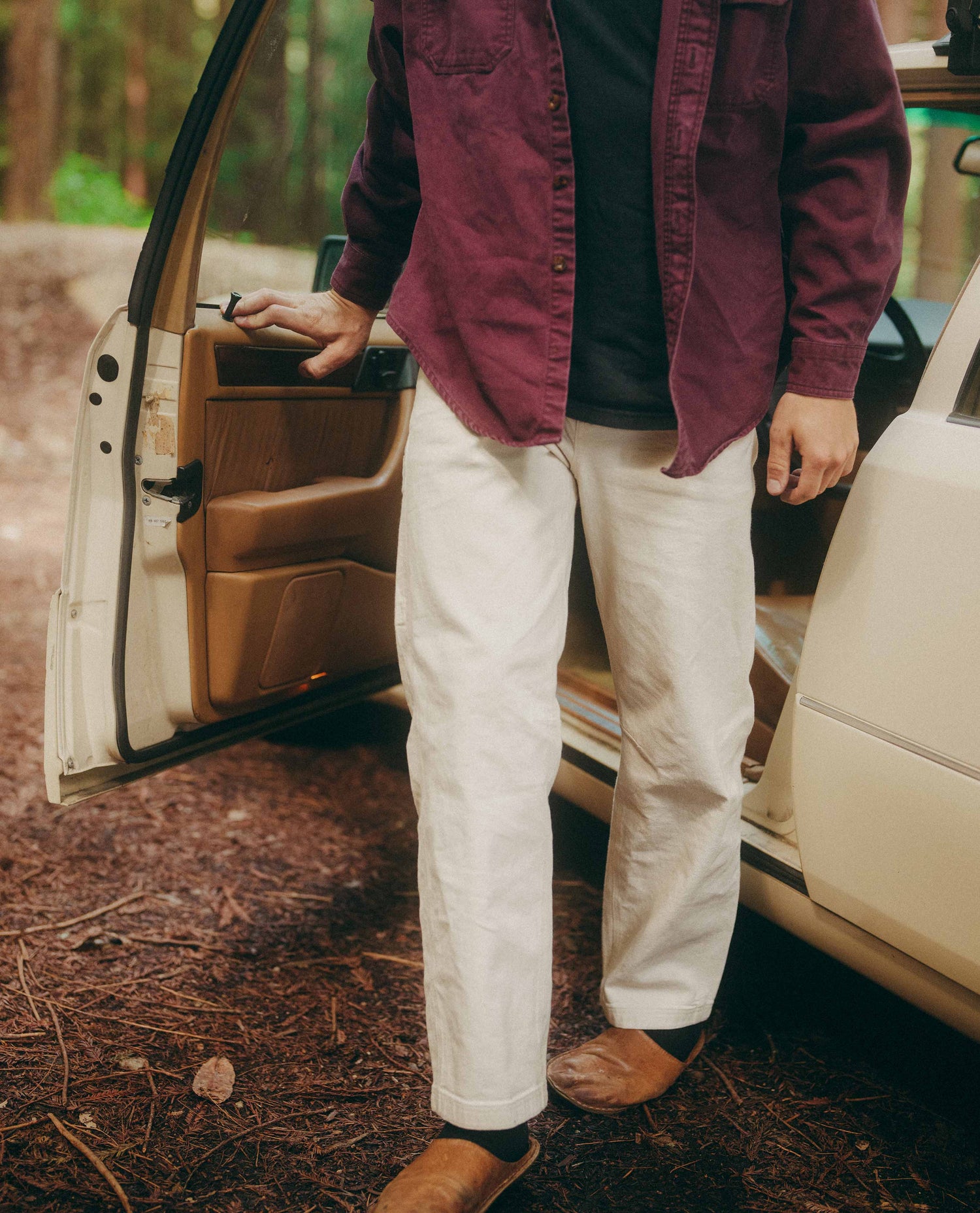 Person in a maroon jacket and white pants standing next to a car in a forest setting