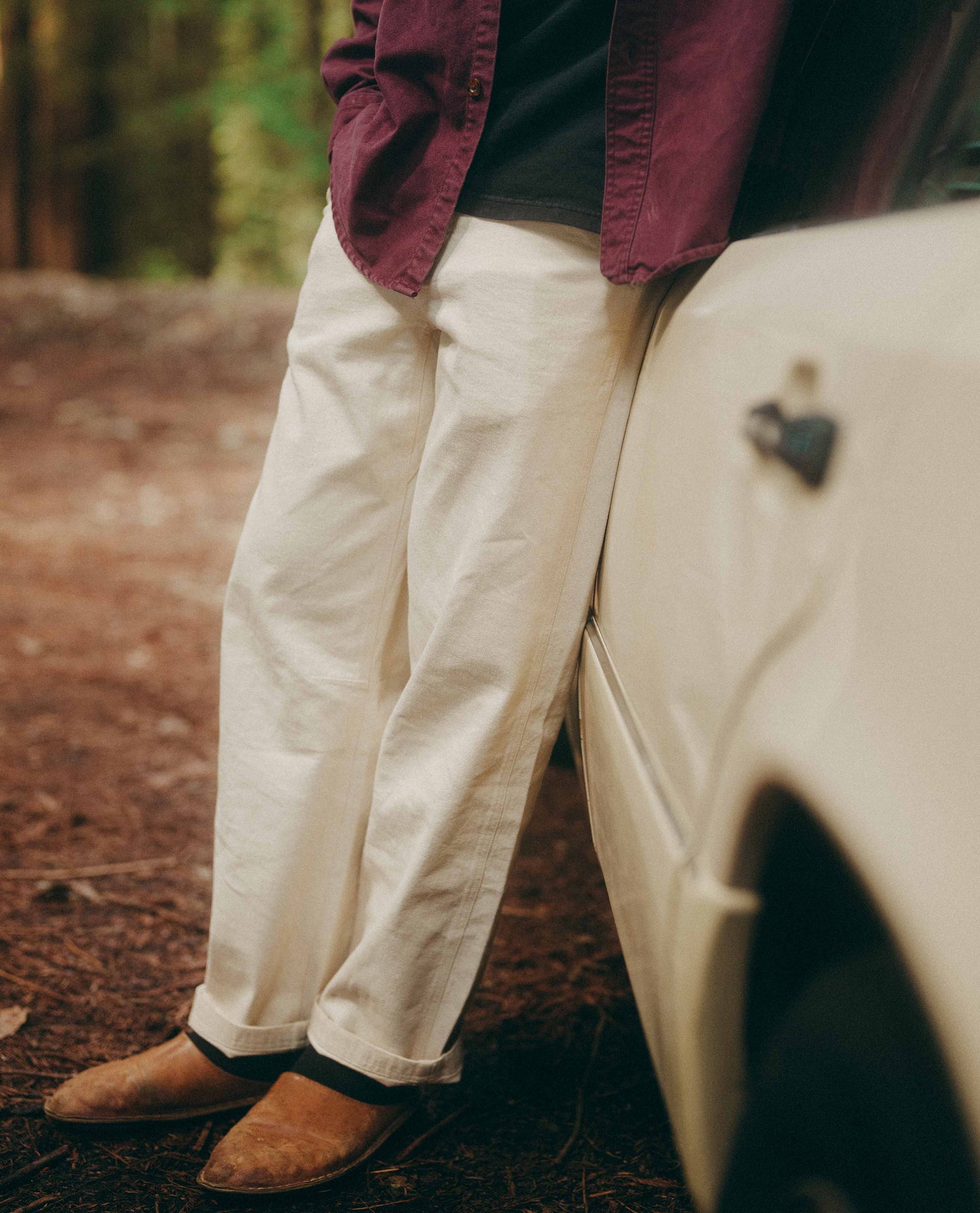 Person wearing a maroon shirt and white pants leaning against a vehicle in a forest setting.