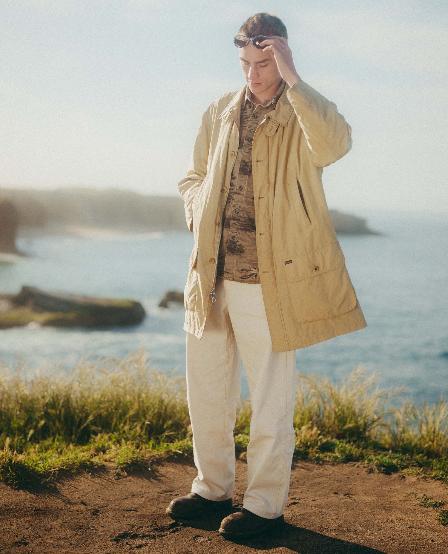 Man standing by a body of water wearing a beige jacket and white pants.