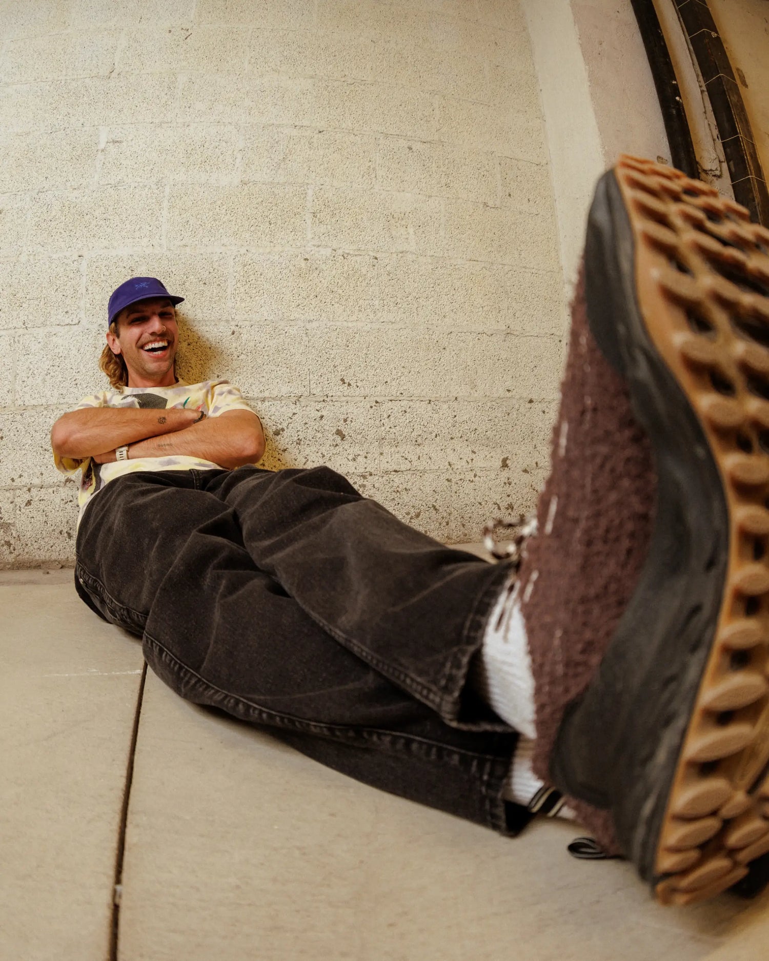 Man leaning against white wall, wearing blue hat, multi-color shirt, black pants, brown shoes