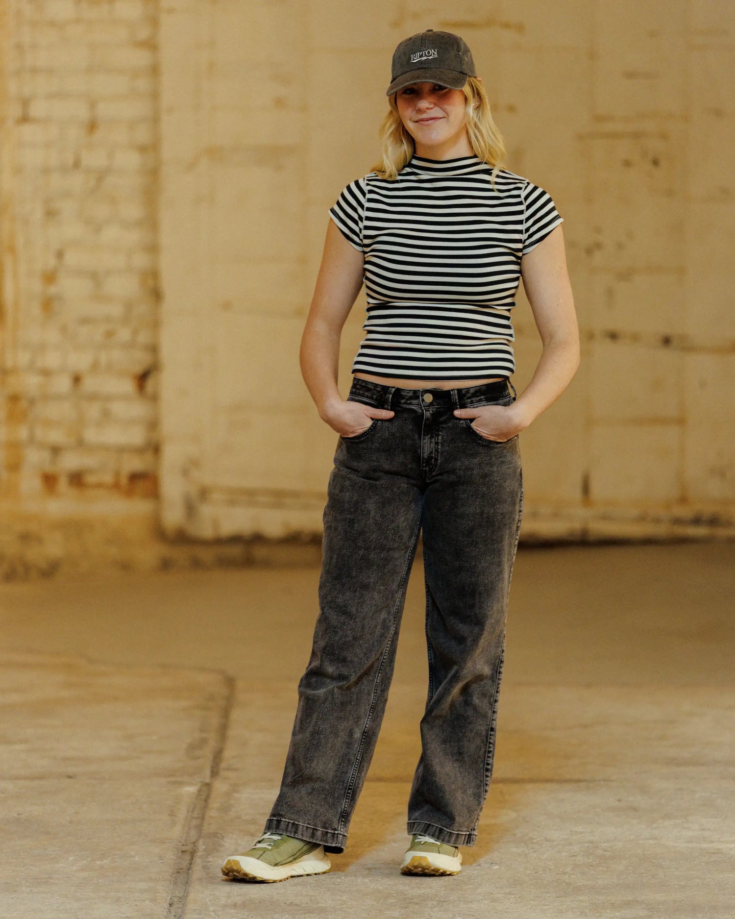 Woman in front of white wall wearing grey hat and pants, striped shirt, and green shoes with hands in pockets 