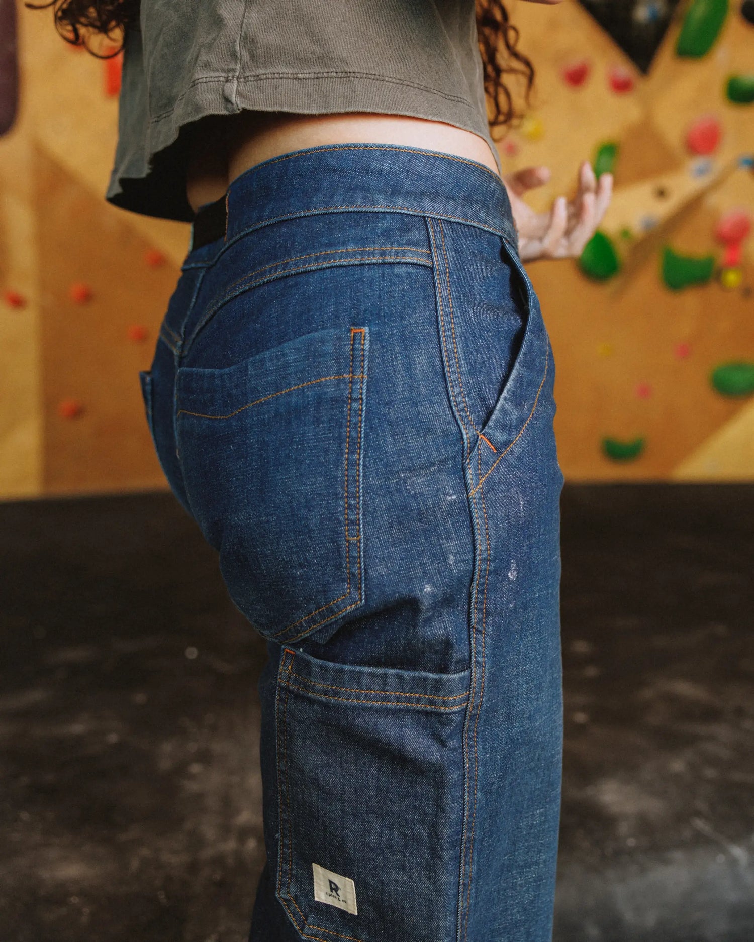 Close up of blue pants with pockets on woman wearing gray shirt in front of climbing gym wall 
