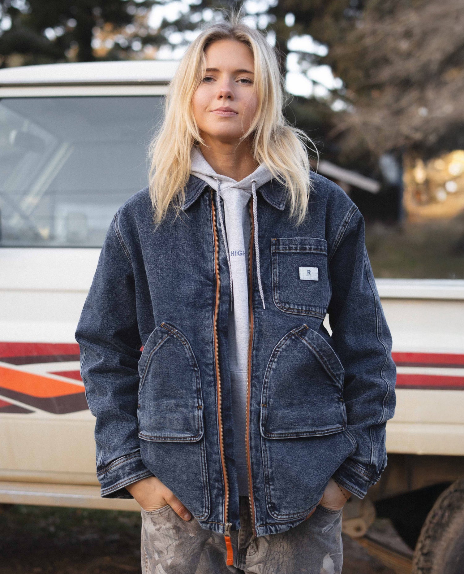 Person wearing a blue denim jacket standing in front of a vehicle with trees in the background Color:Dark