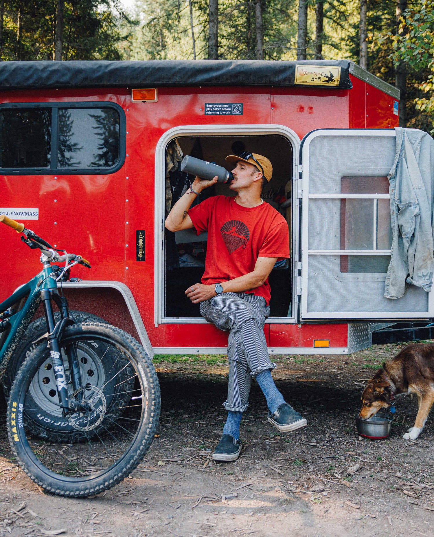 A man in a red graphic t-shirt and black and white striped pants sits on the open doorway of a red camping trailer, drinking from a water bottle. A dog is eating from a bowl on the ground nearby, and a mountain bike is parked on the left, all set in a forest environment.