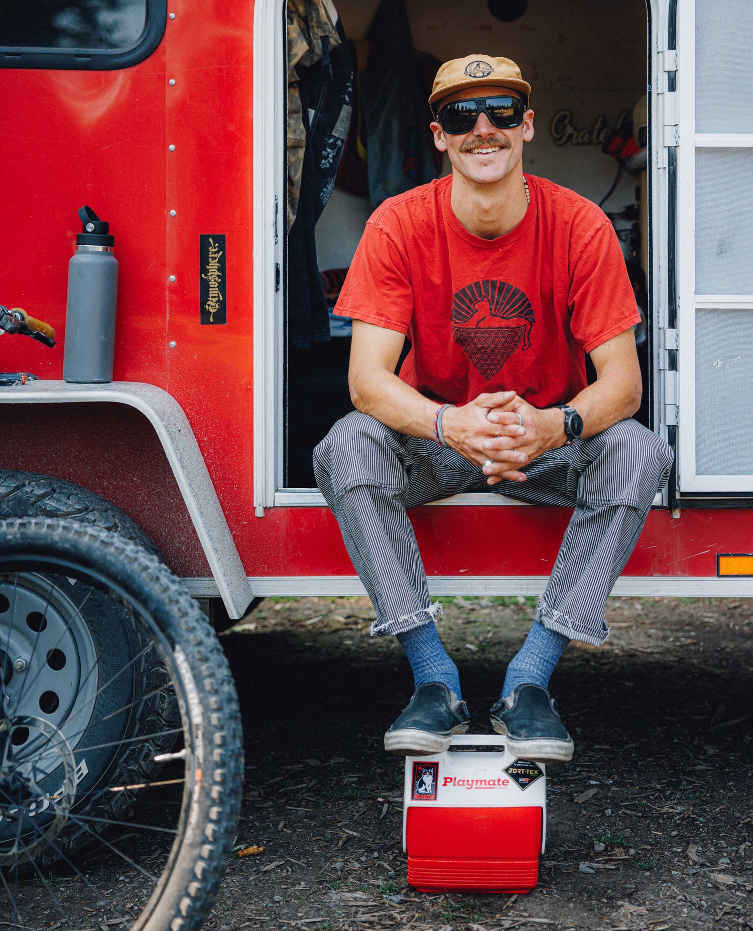 A man wearing a red graphic t-shirt, gray jeans, and a tan cap sits on the step of a red trailer next to a bicycle tire, with a red and white cooler in front of him.