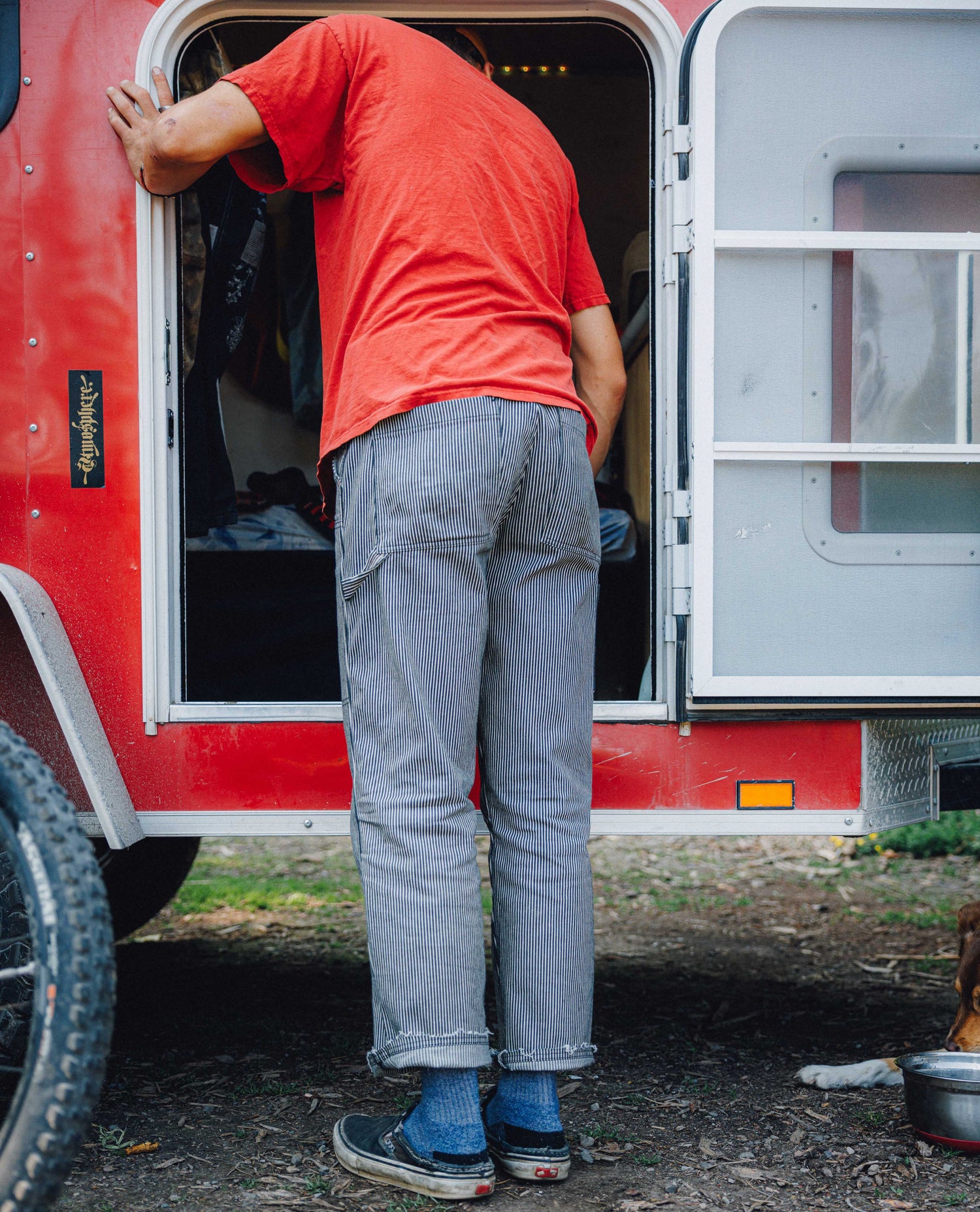 A man stands outside a red camper. He's wearing a red shirt and black and white striped pants with blue socks and black shoes. 